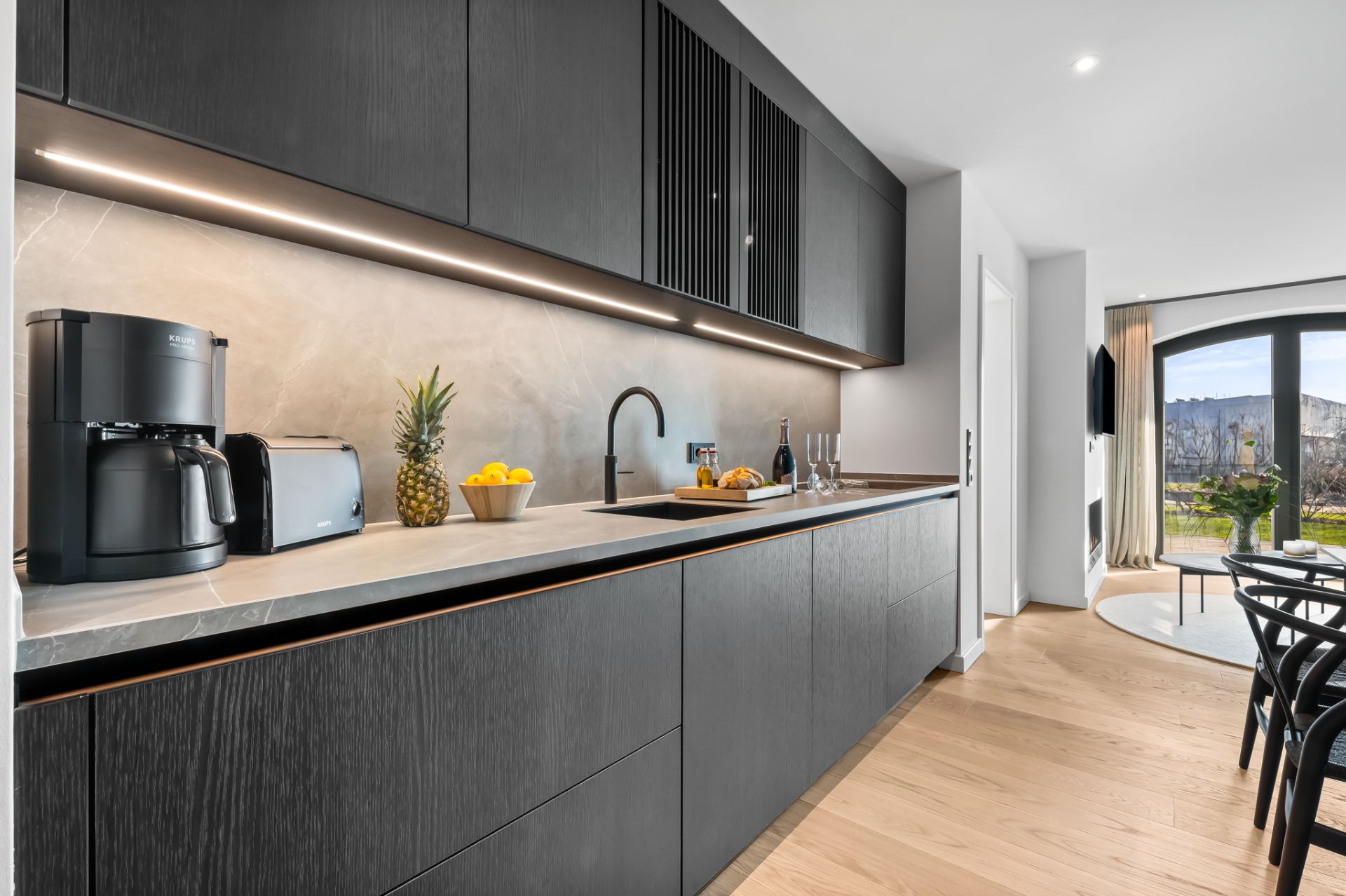 Moderne Küche mit schwarzen Schränken, Kaffeemaschine und Obst, daneben heller Wohnbereich mit großem Fenster.