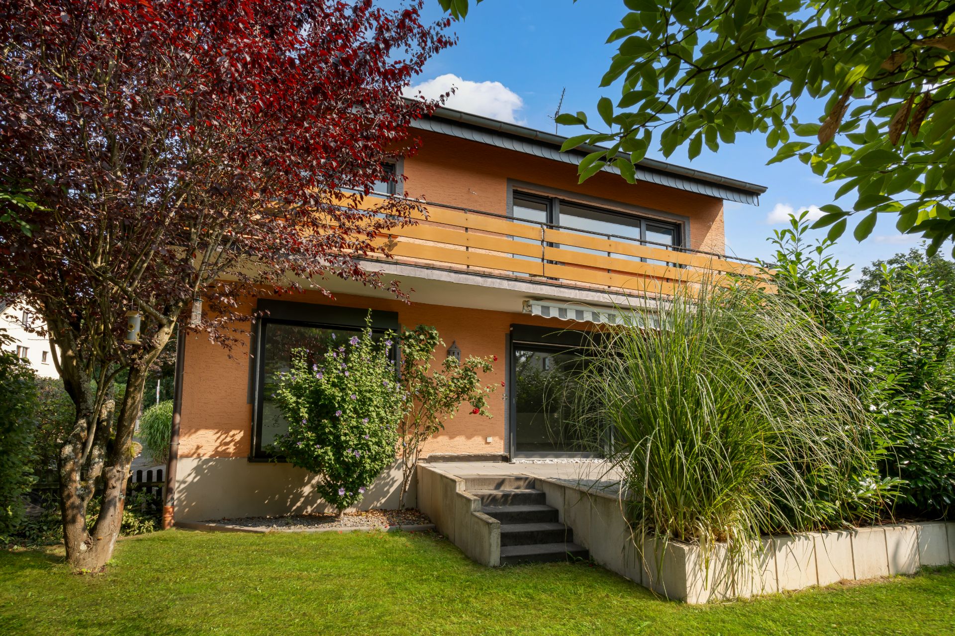 Zweistöckiges Wohnhaus mit Balkon, Terrasse und Garten, umgeben von Bäumen und Sträuchern.