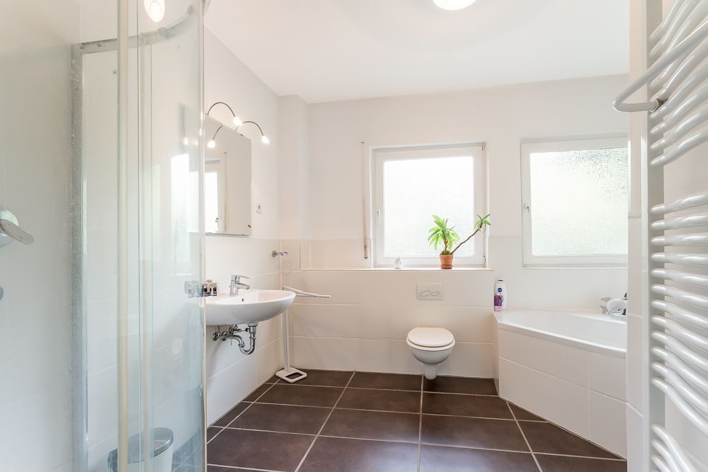 Helles Badezimmer mit Dusche, Waschbecken, Toilette, Badewanne und zwei Fenstern.