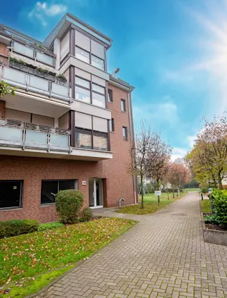 Mehrfamilienhaus mit Balkonen, Vorgarten und Gehweg bei sonnigem Wetter.
