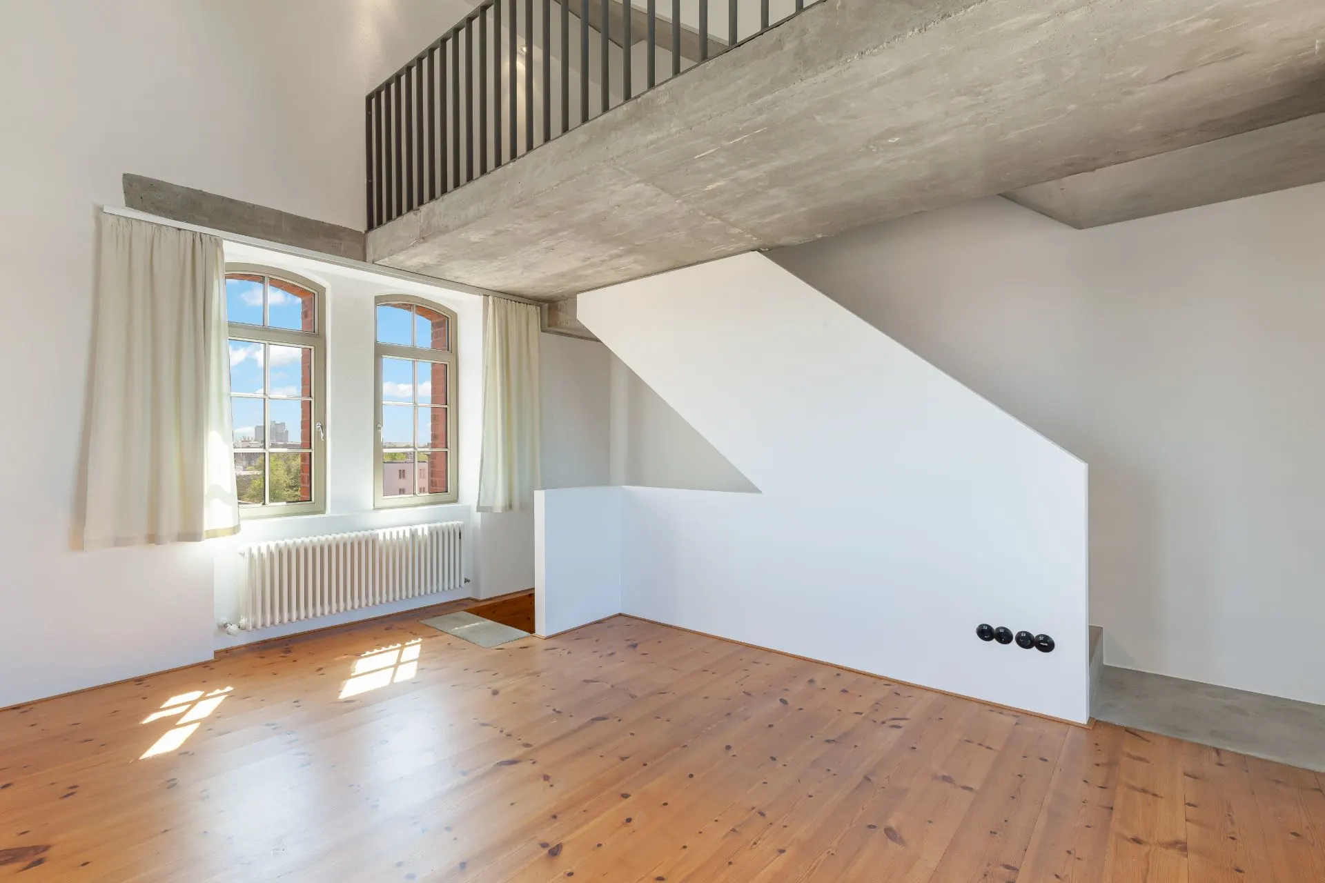 Helles Zimmer mit Holzfußboden, großen Fenstern, Heizung und einer offenen Treppe zu einer Galerie.