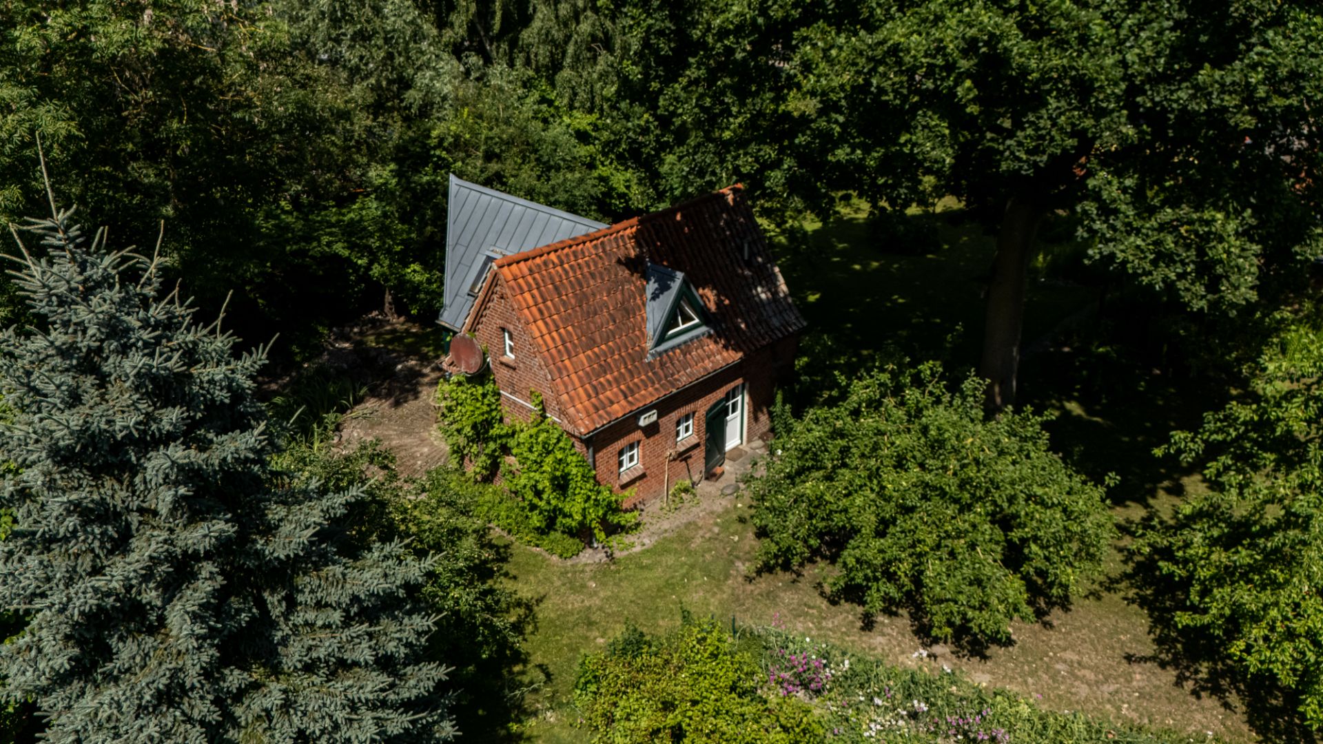 Kleines Backsteinhaus mit rotem Dach, umgeben von Bäumen und Garten aus der Vogelperspektive.