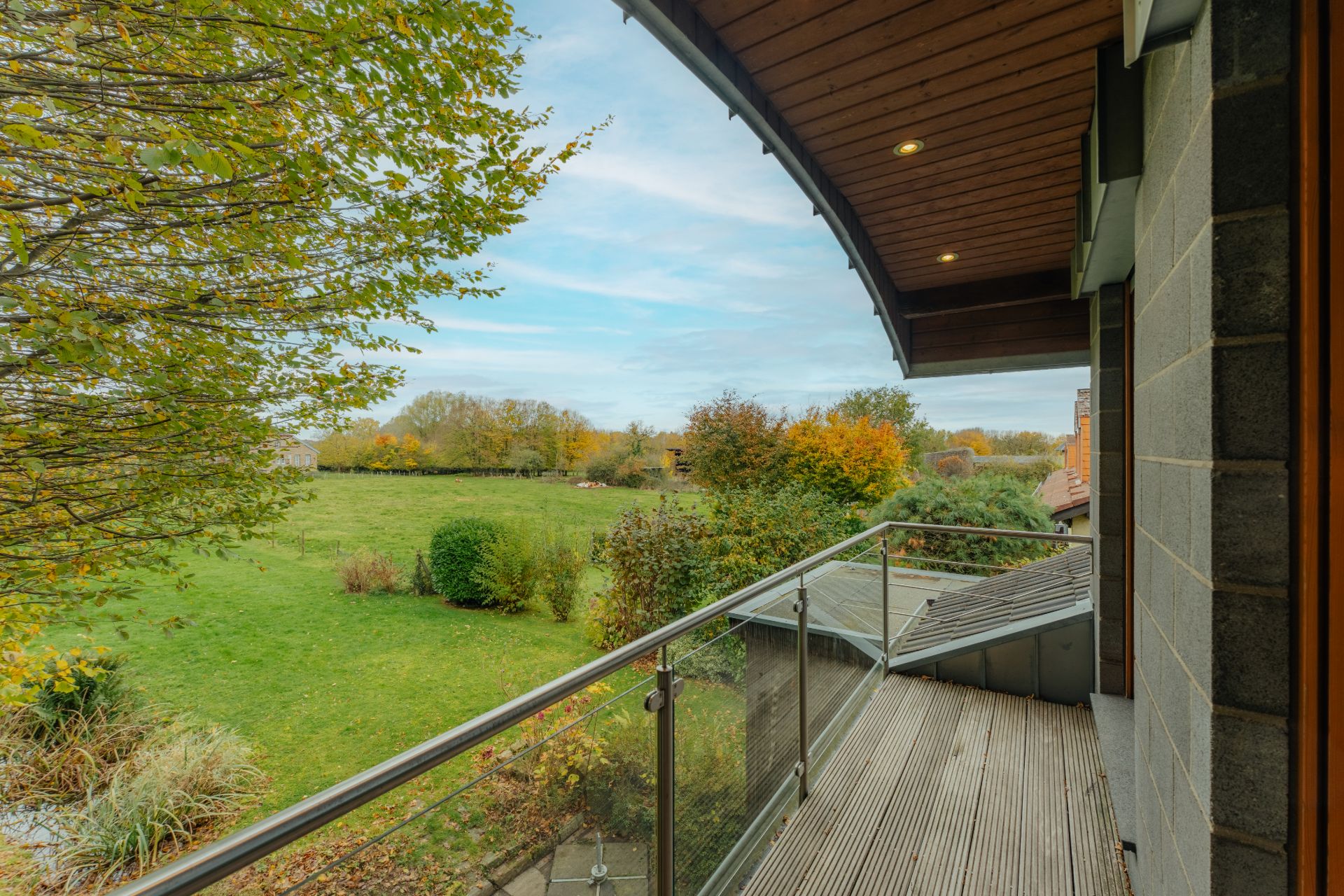 Balkon mit Glasgeländer und Blick auf einen Garten mit Bäumen und Wiese.