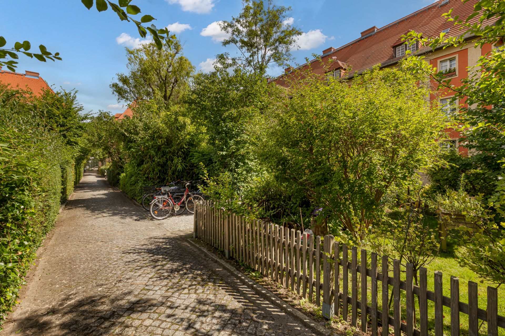 Gepflasterter Weg mit zwei Fahrrädern, umgeben von Bäumen und Wohnhäusern im Hintergrund.