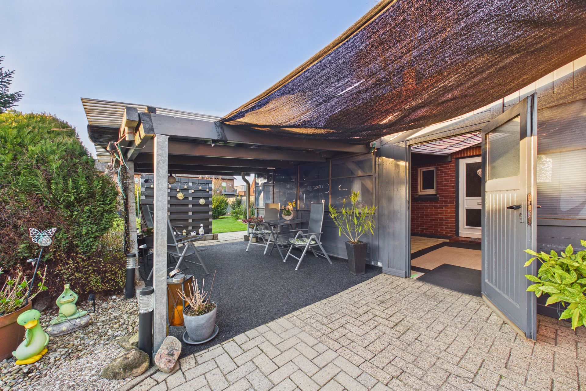 Überdachte Terrasse mit Gartenmöbeln, Pflanzen und Blick auf einen Gartenbereich.