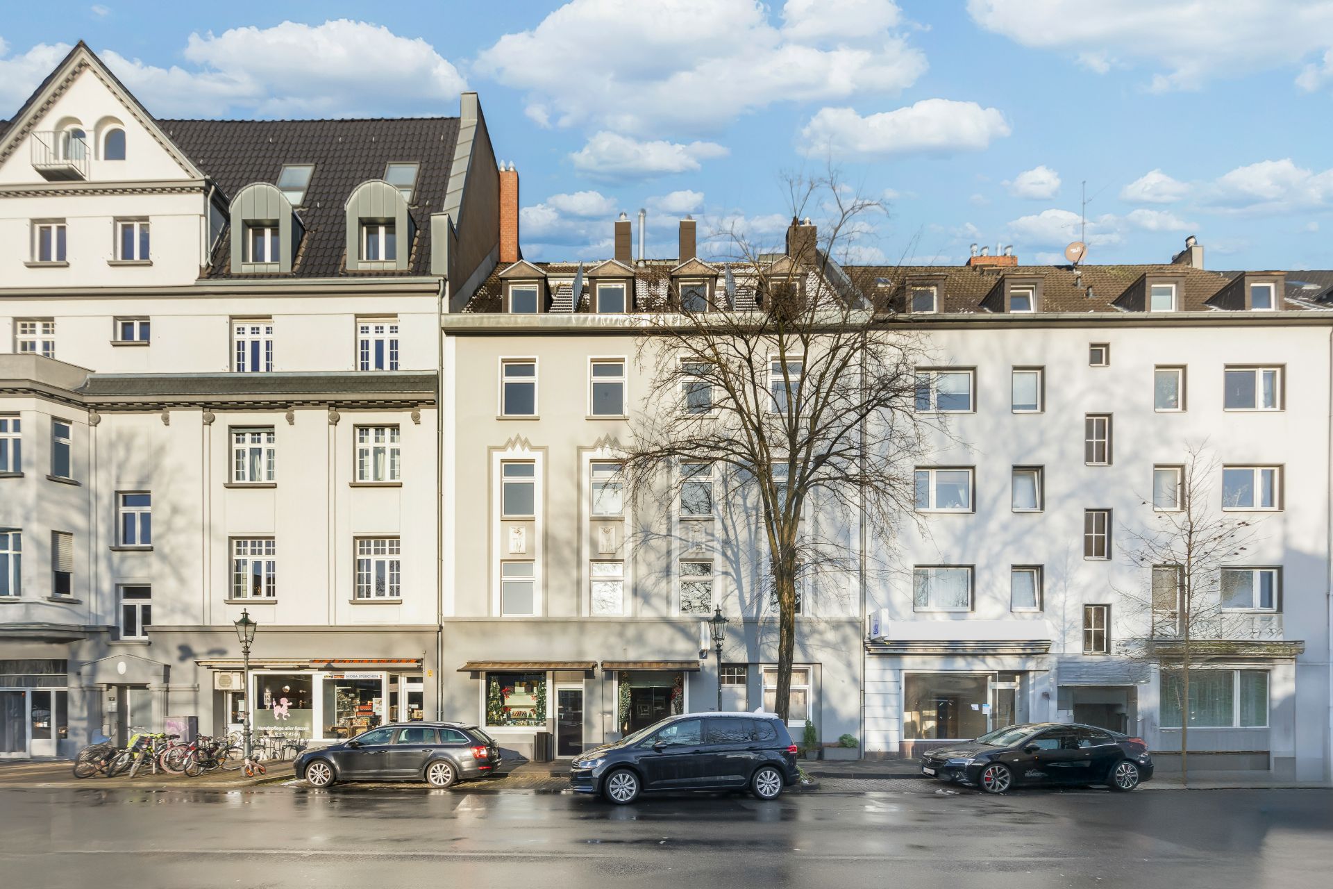 Wohnhäuser mit Schaufenstern im Erdgeschoss, geparkte Autos und Fahrräder davor.
