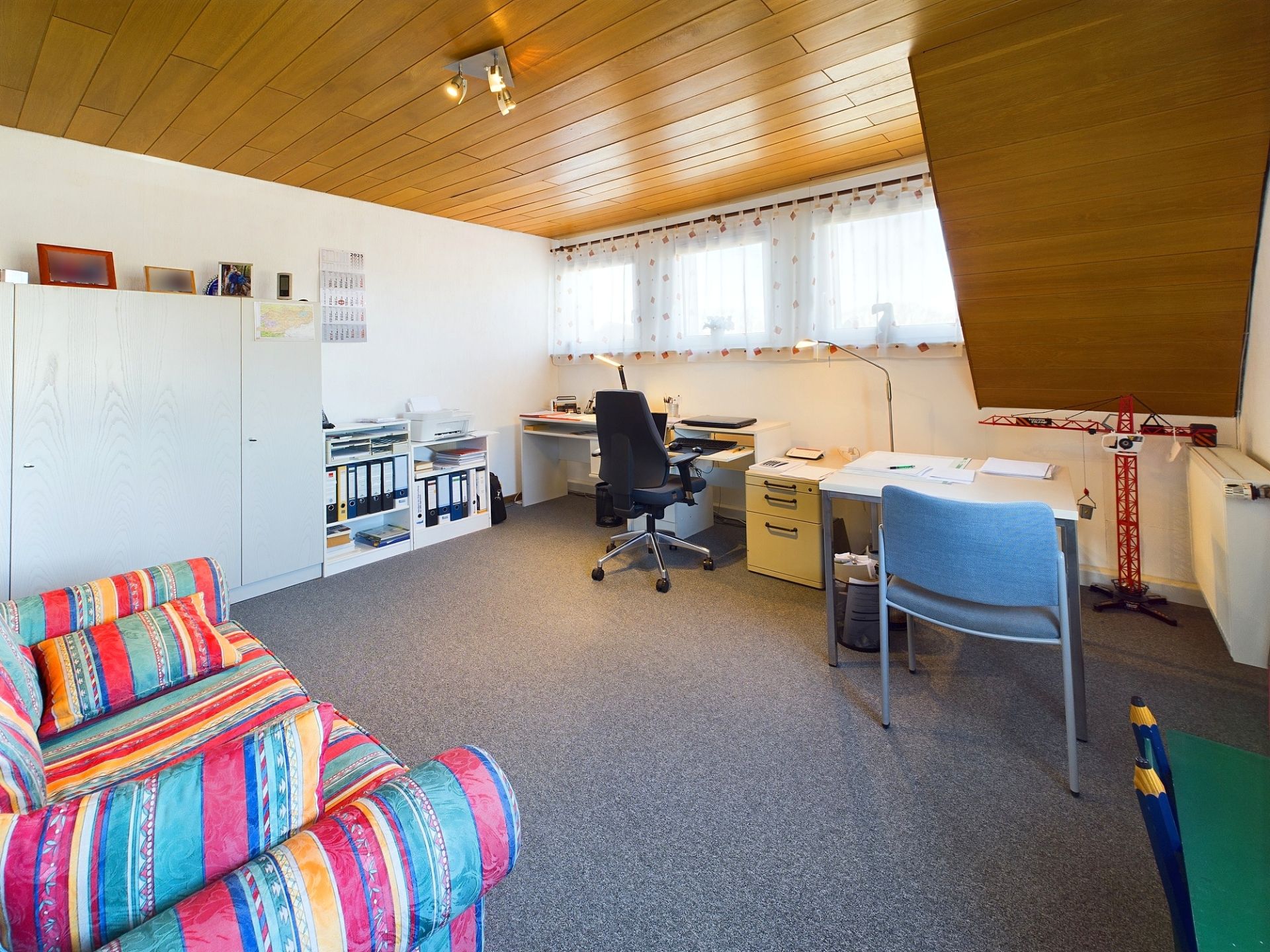 Helles Arbeitszimmer mit zwei Schreibtischen, Regalen, Schränken und bunter Couch.