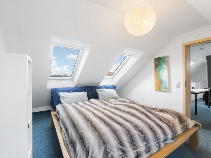 Schlafzimmer mit Doppelbett unter zwei Dachfenstern, blauer Teppichboden und weißem Regal.