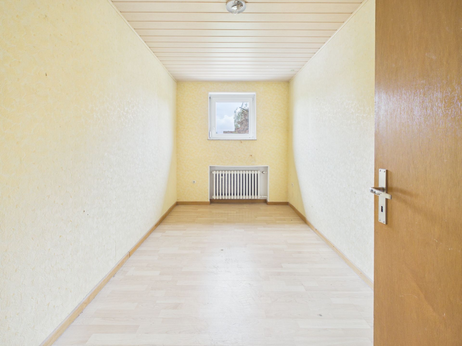 Leerer, schmaler Raum mit gelben Wänden, Fenster und Heizkörper am Ende, Holztür offen.