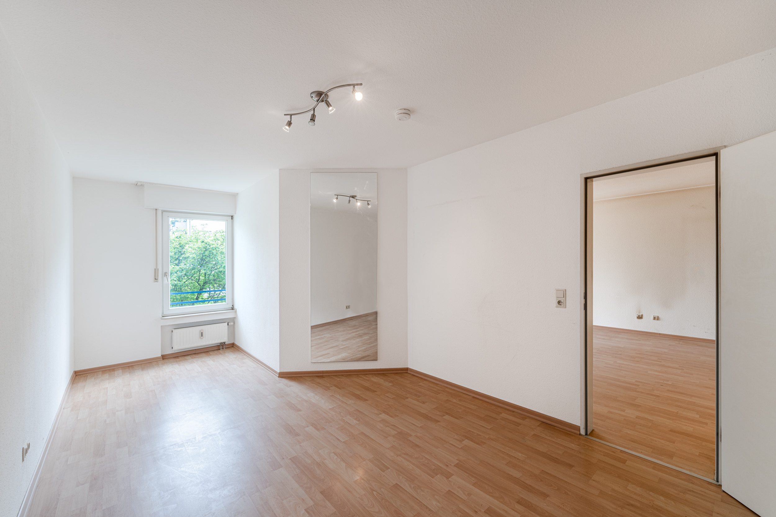 Leeres Zimmer mit Holzfußboden, Fenster, Spiegel und offenem Durchgang zu weiterem Raum.