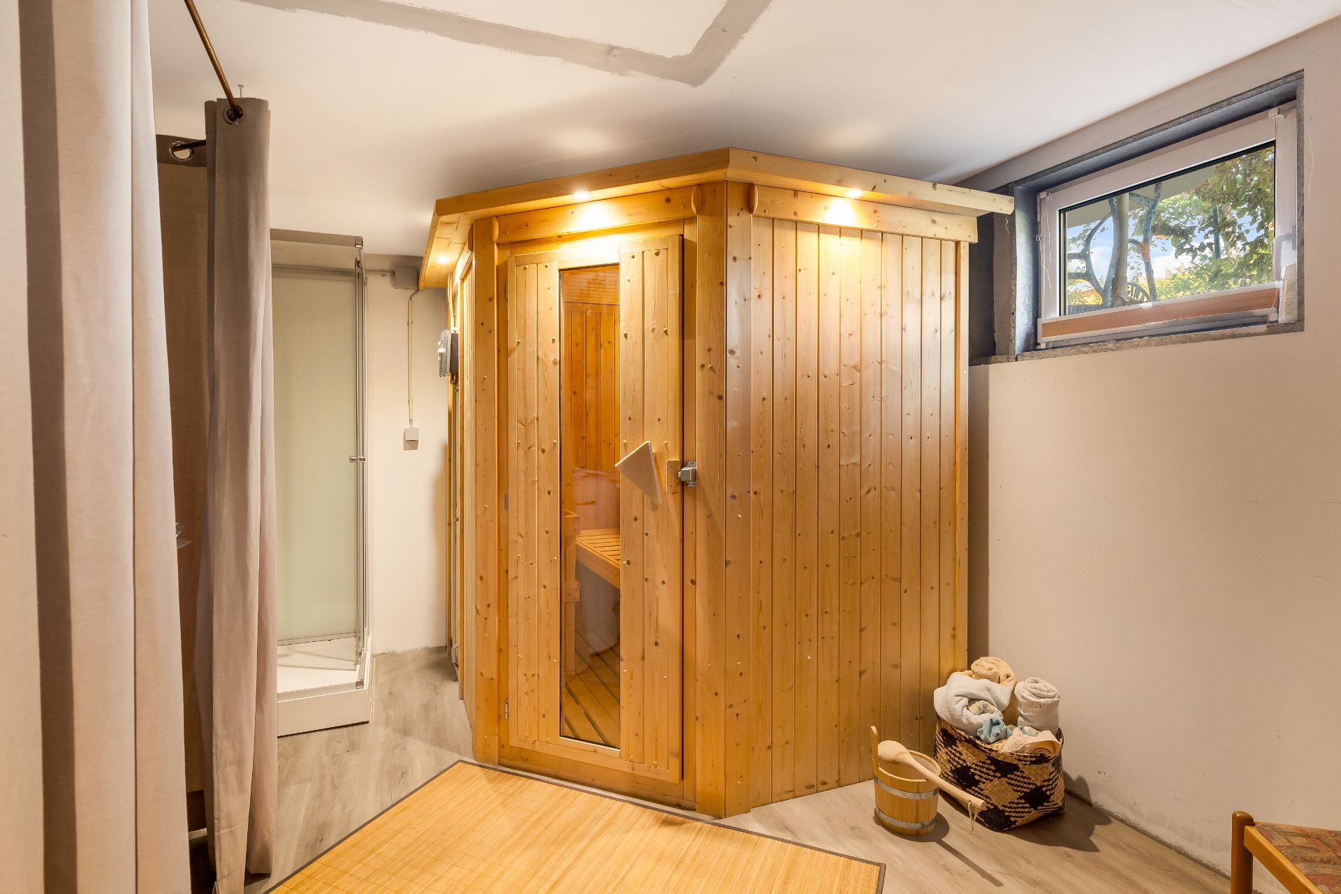 Holzsauna mit Glastür, daneben Dusche, Korb mit Handtüchern und Fenster im Raum.