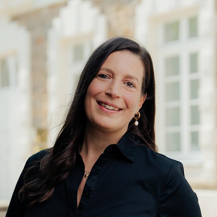 Frau mit langen dunklen Haaren und schwarzer Bluse steht vor einem Fenster.