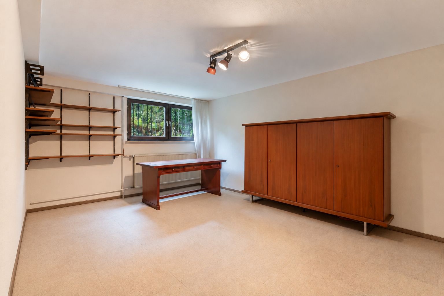 Raum mit Schreibtisch, Holzschrank, Regal an der Wand und Fenster mit Blick ins Grüne.