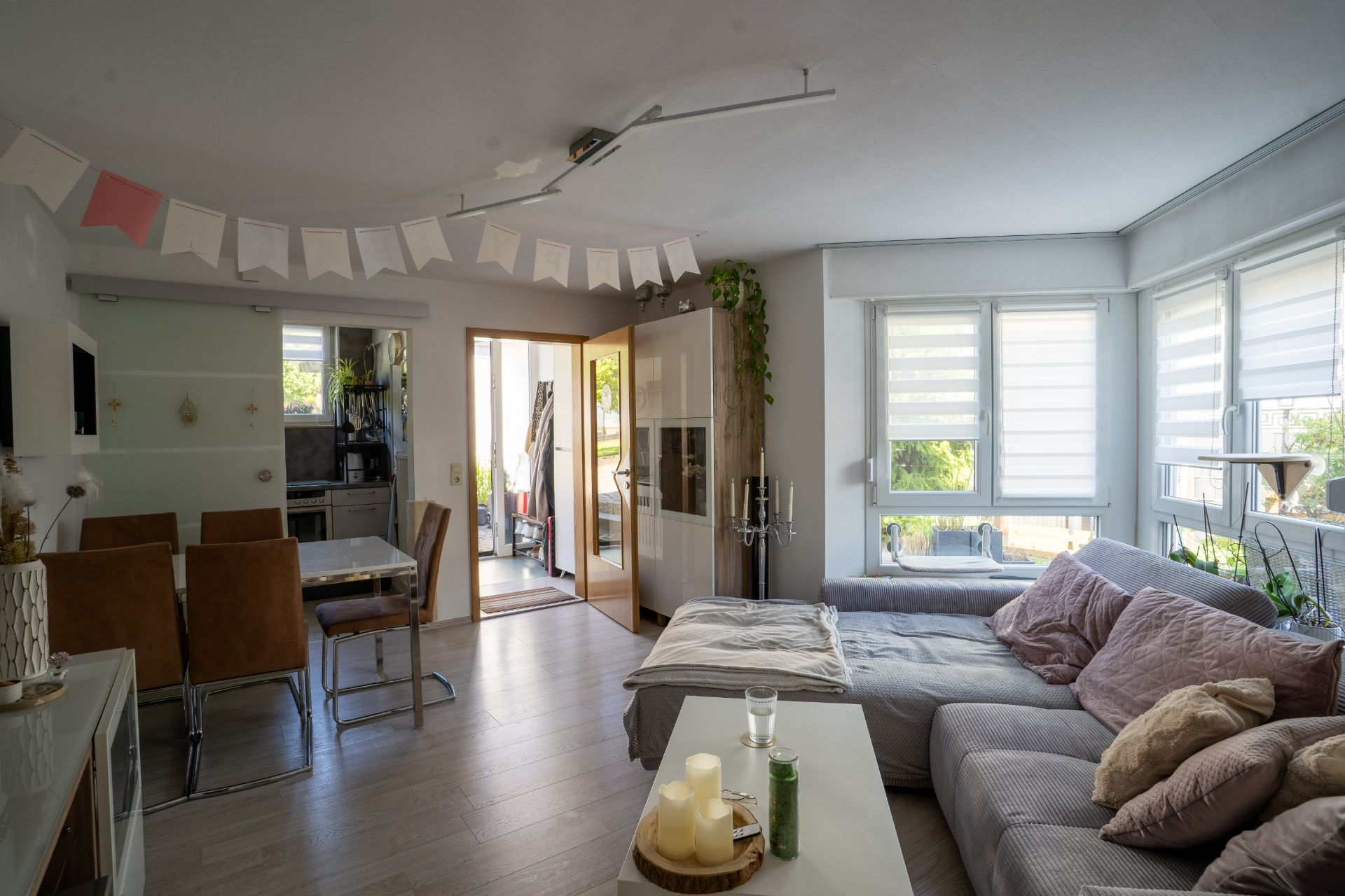 Helles Wohnzimmer mit Ecksofa, Esstisch, großen Fenstern und Dekorationen.