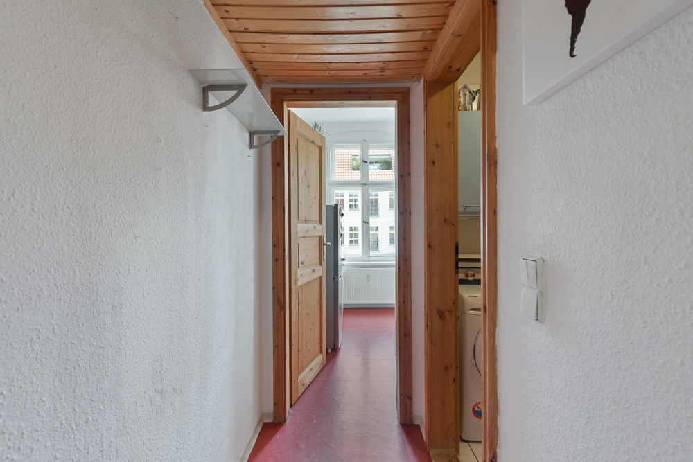 Schmaler Flur mit Holzdecke, rotem Boden und Blick in einen Raum mit großen Fenstern.