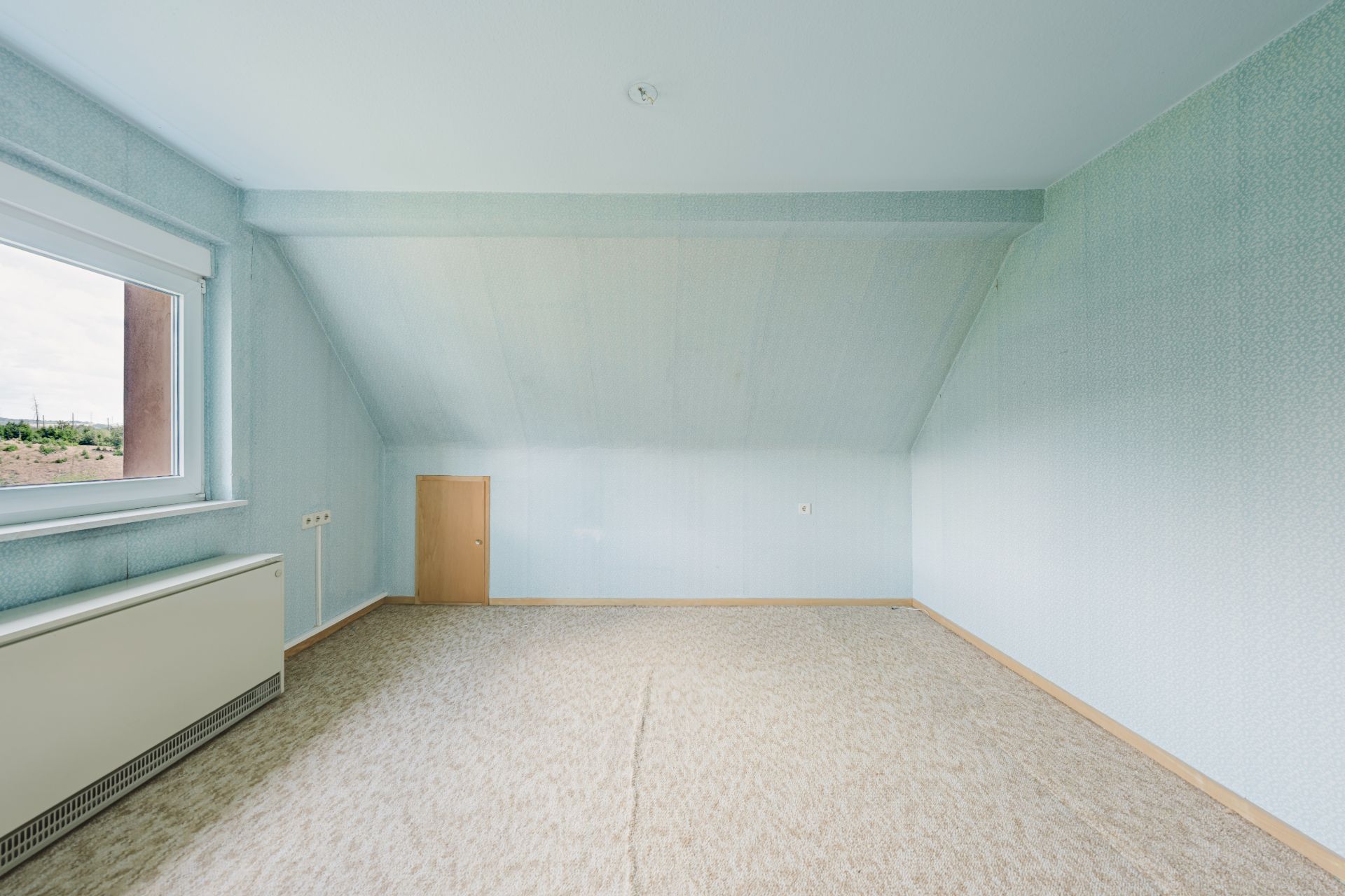 Leerer Raum mit schräger Decke, Fenster links und kleinem Türchen in der Wand.