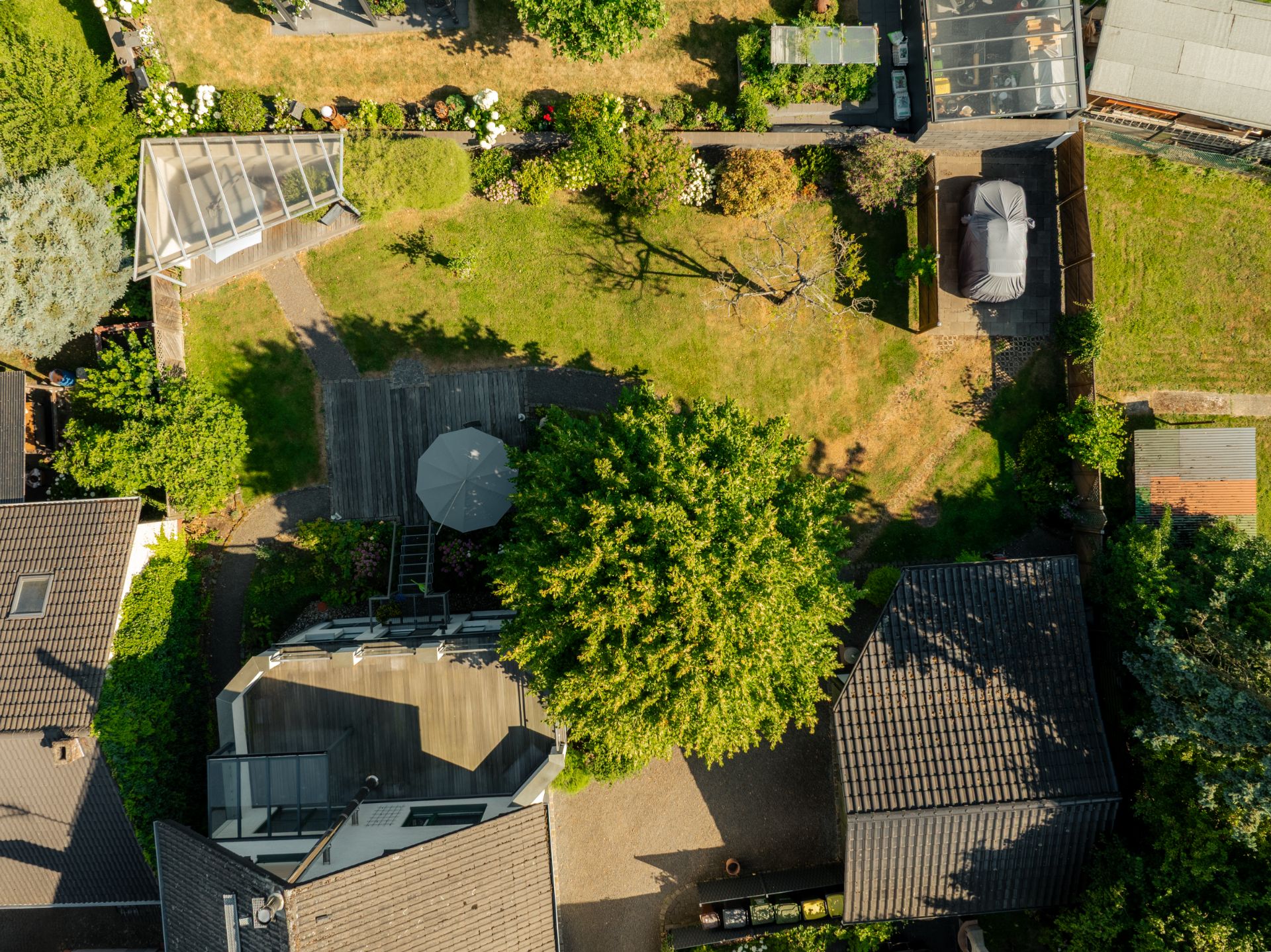 Luftaufnahme eines Gartens mit Terrasse, Sonnenschirm, Bäumen und umliegenden Wohnhäusern.