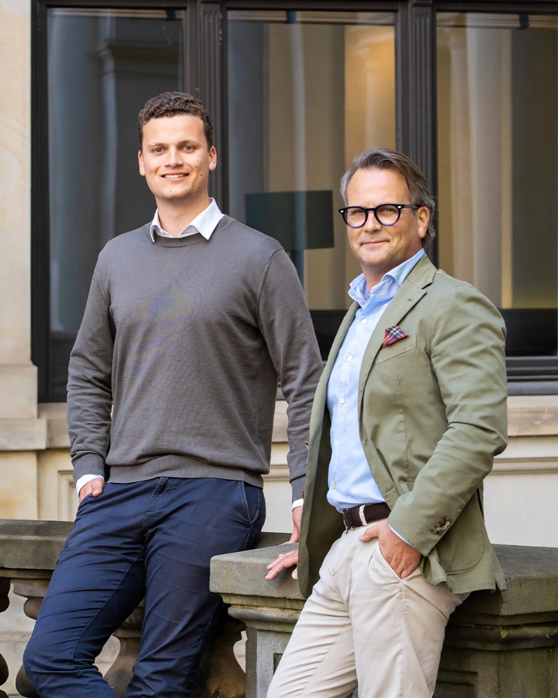 Zwei Männer stehen lächelnd an einer Steintreppe vor einem großen Fenster.