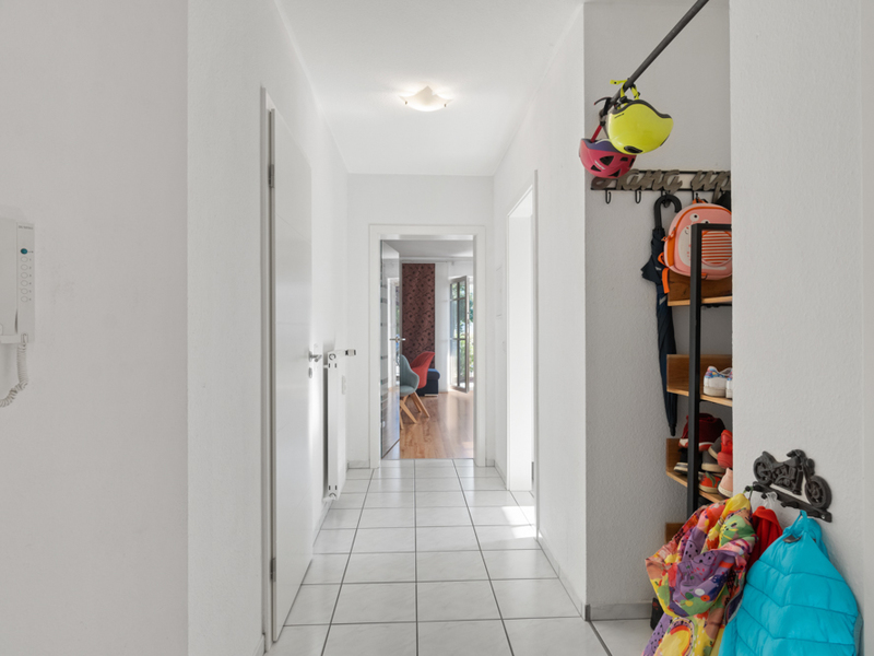 Heller Flur mit weißen Wänden, Garderobe und Fliesenboden, Tür zum Wohnzimmer im Hintergrund.
