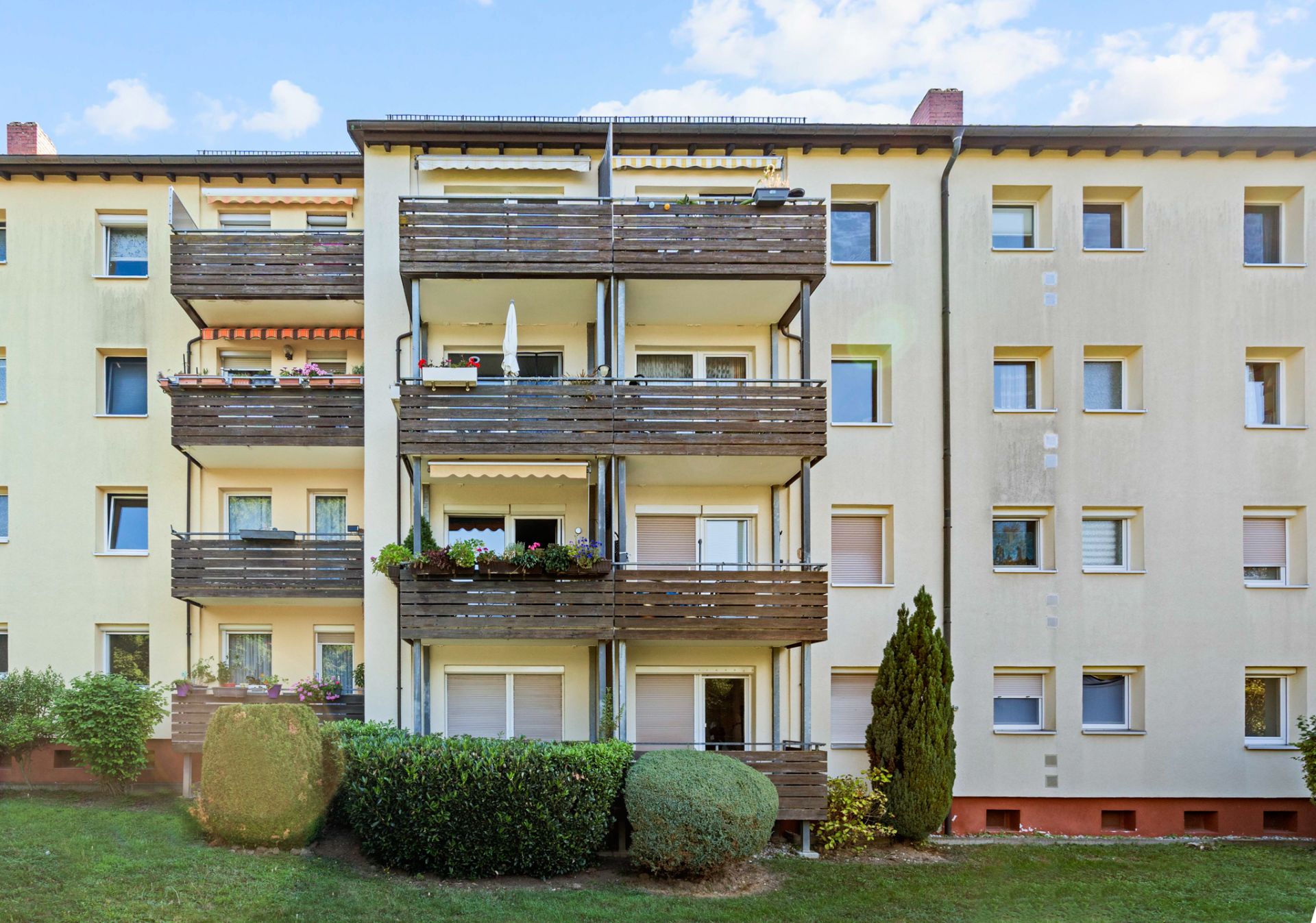 Mehrfamilienhaus mit drei Etagen, Balkonen und gepflegten Büschen im Vordergrund.