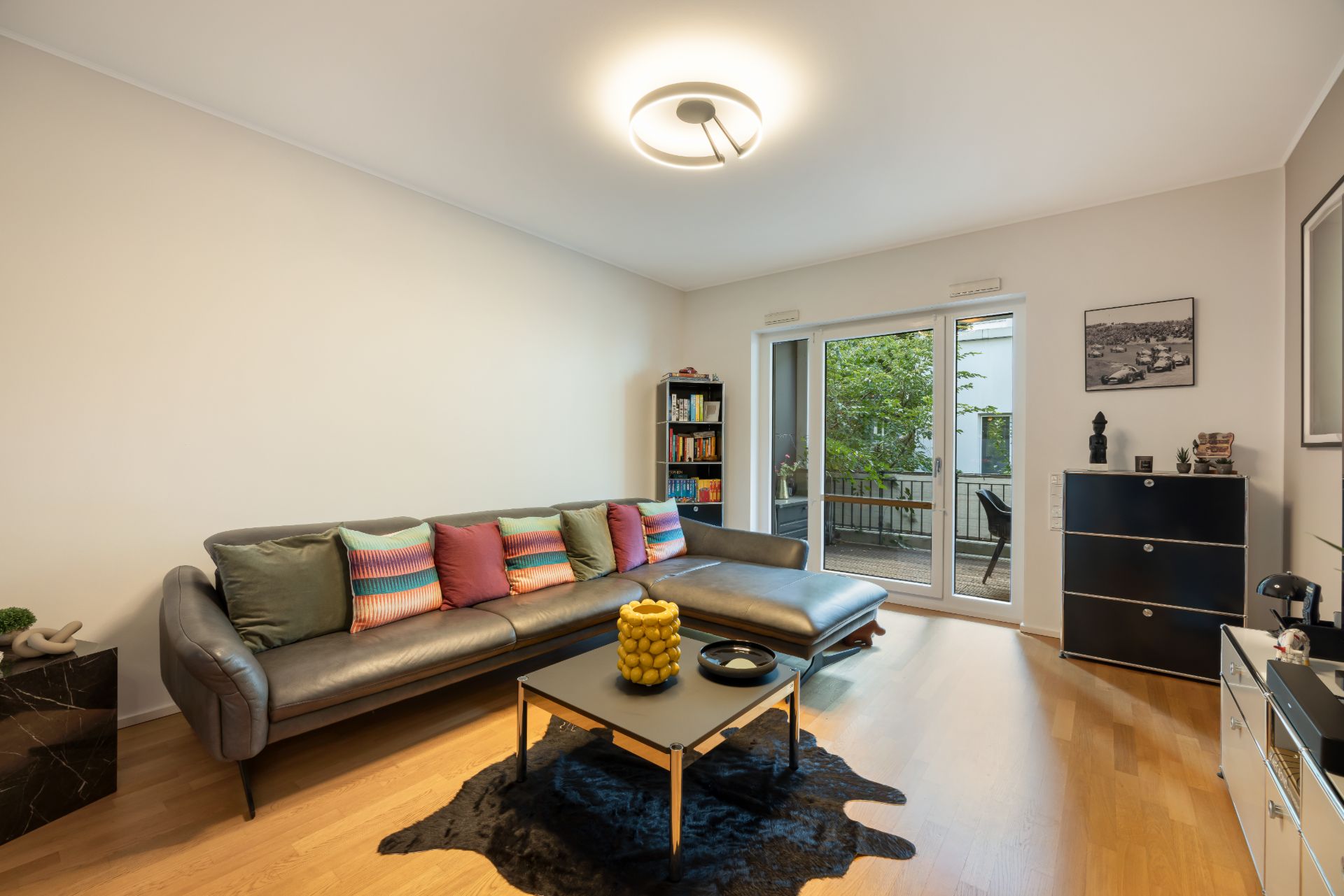 Modernes Wohnzimmer mit Ecksofa, Couchtisch, Bücherregal und Zugang zu einem Balkon mit Blick ins Grüne.