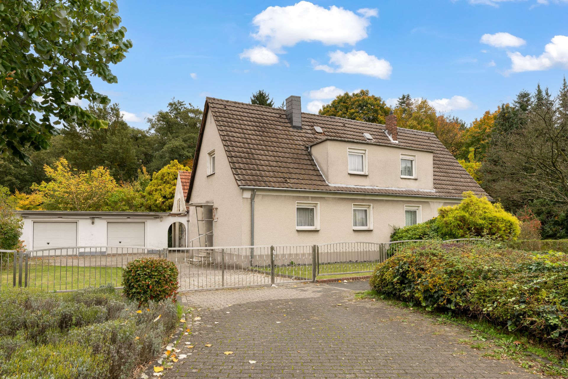 Zweistöckiges Wohnhaus mit Zaun, Einfahrt und angrenzender Garage, umgeben von Bäumen und Büschen.