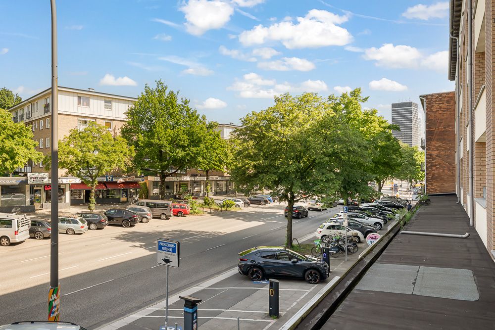 Straßenszene mit parkenden Autos, Bäumen und einem Fußweg vor Wohn- und Geschäftsgebäuden.