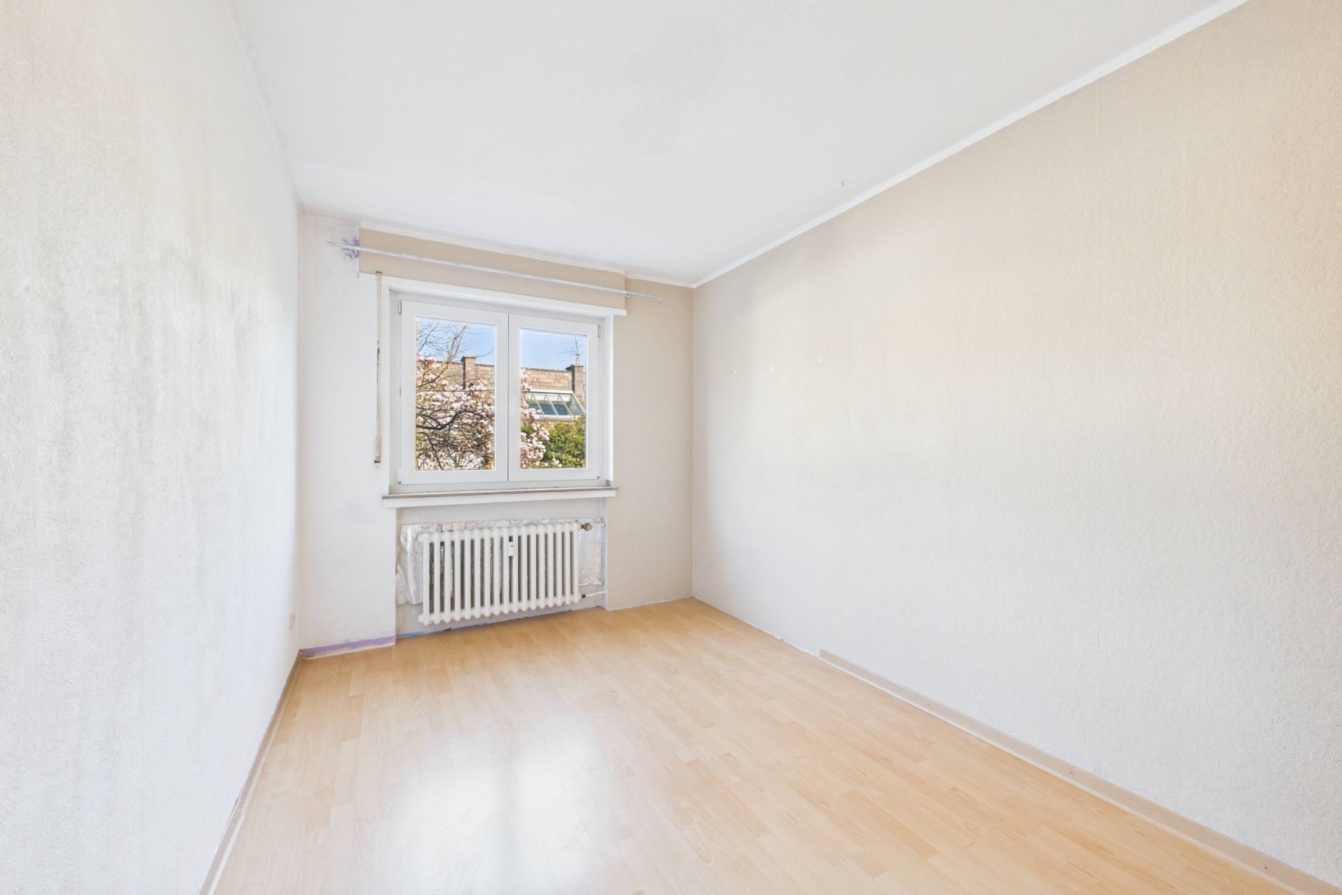 Leerer Raum mit hellem Laminatboden, weißer Heizung und Fenster mit Blick nach draußen.