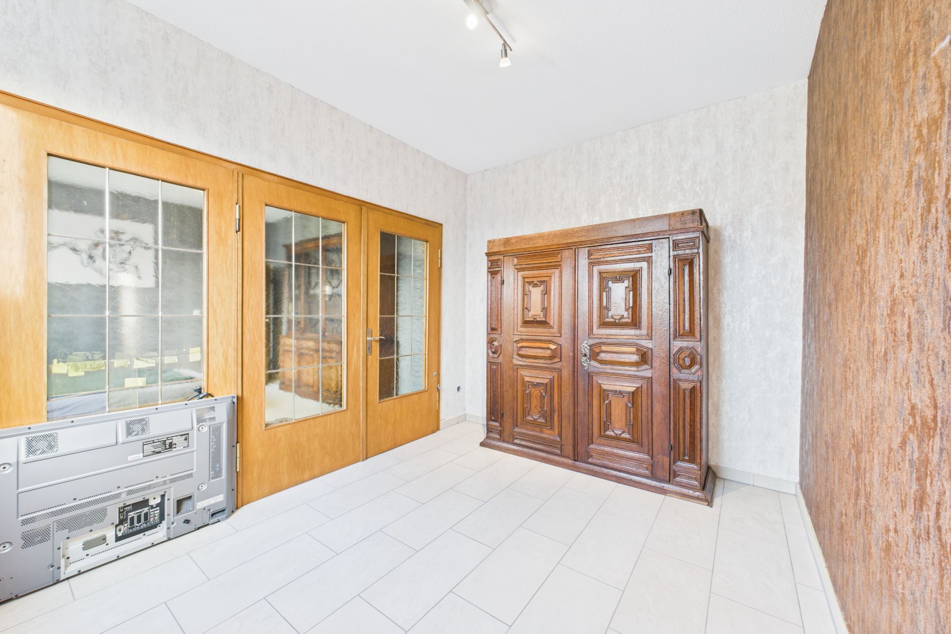 Raum mit großem Holzschrank, Glastüren und einem alten Fernseher auf weißem Fliesenboden.
