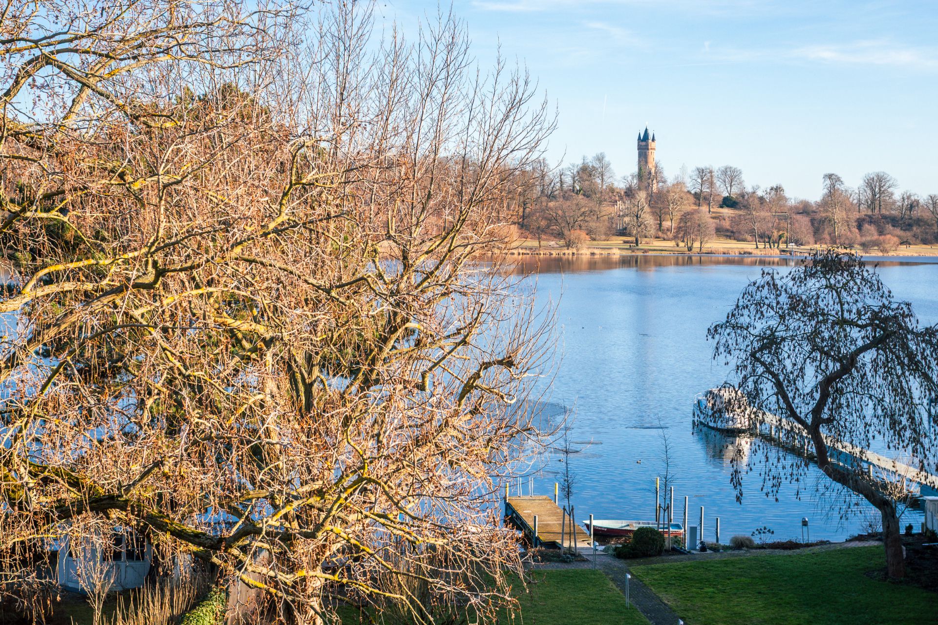 Blick auf einen See mit Steg und Bäumen im Vordergrund, Turm im Hintergrund.