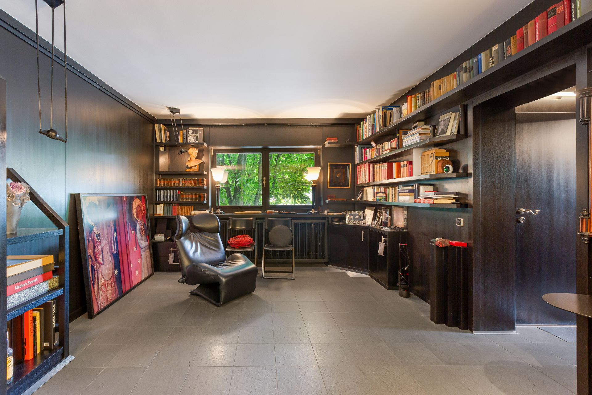 Wohnzimmer mit schwarzen Bücherregalen, Sessel, Bildern an der Wand und großem Fenster.