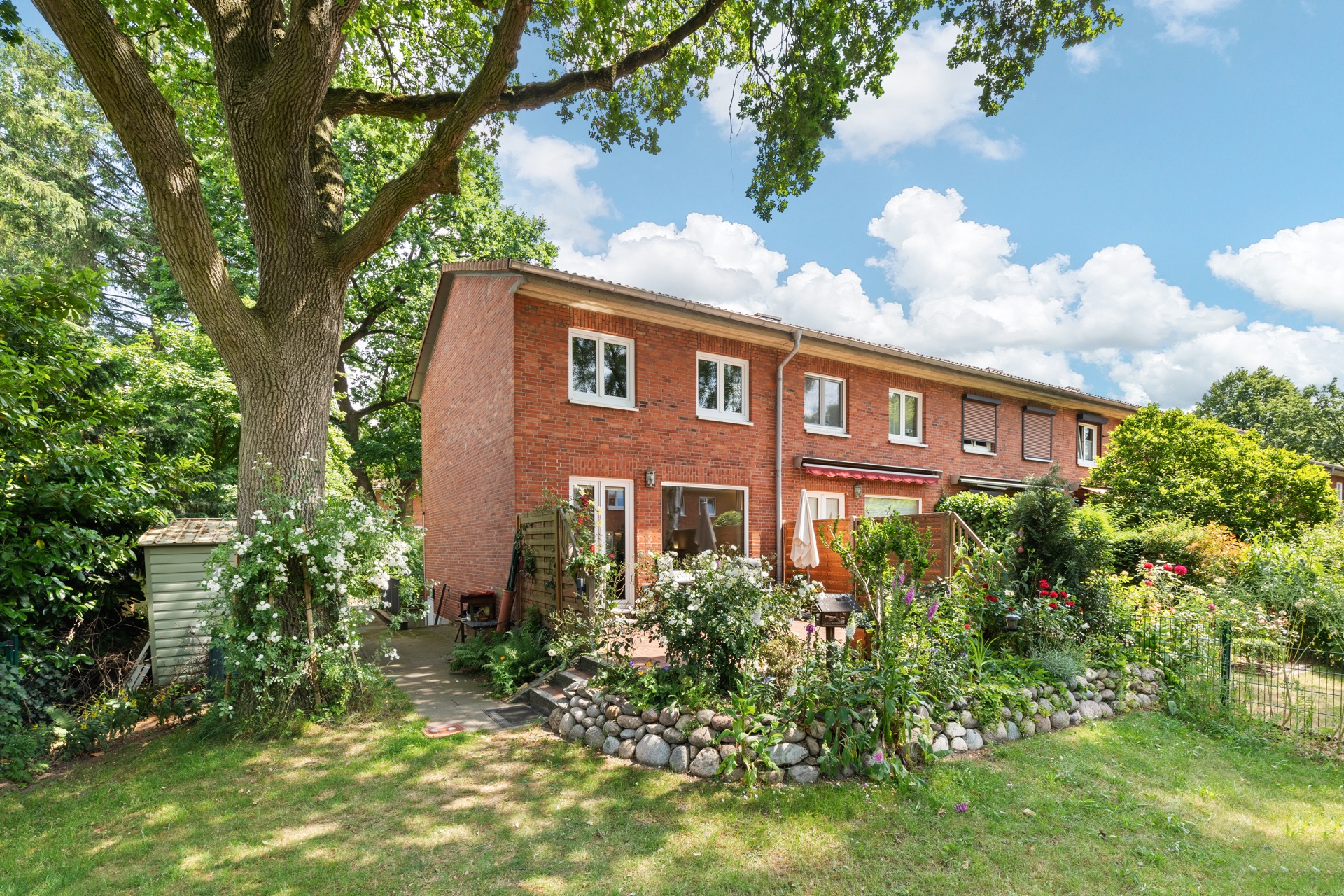 Backstein-Reihenhaus mit Garten, großem Baum und Terrasse, umgeben von viel Grün.
