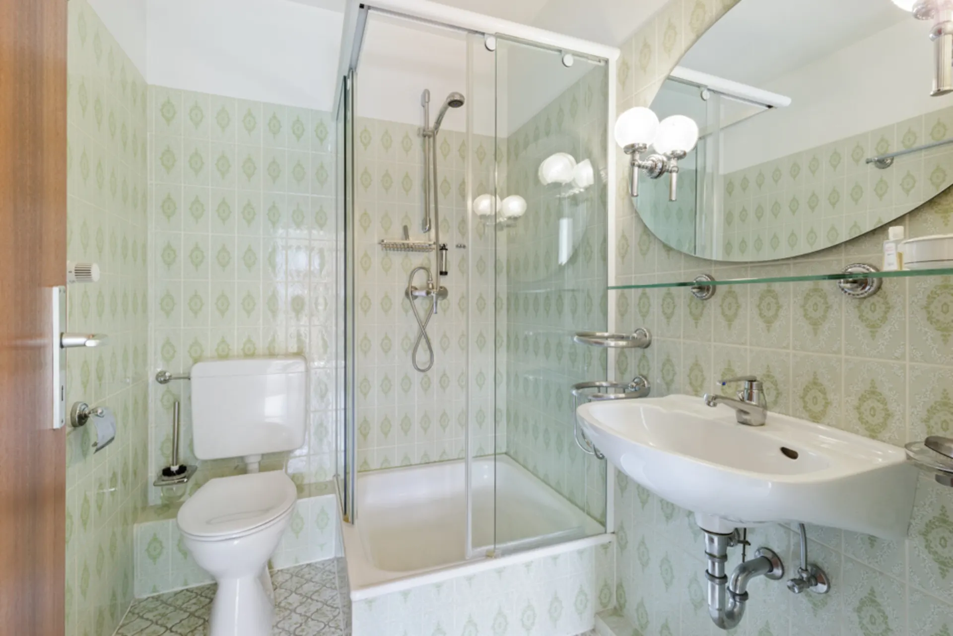Kleines Bad mit Dusche, Toilette, Waschbecken und großem Spiegel, grüne Fliesen an den Wänden.