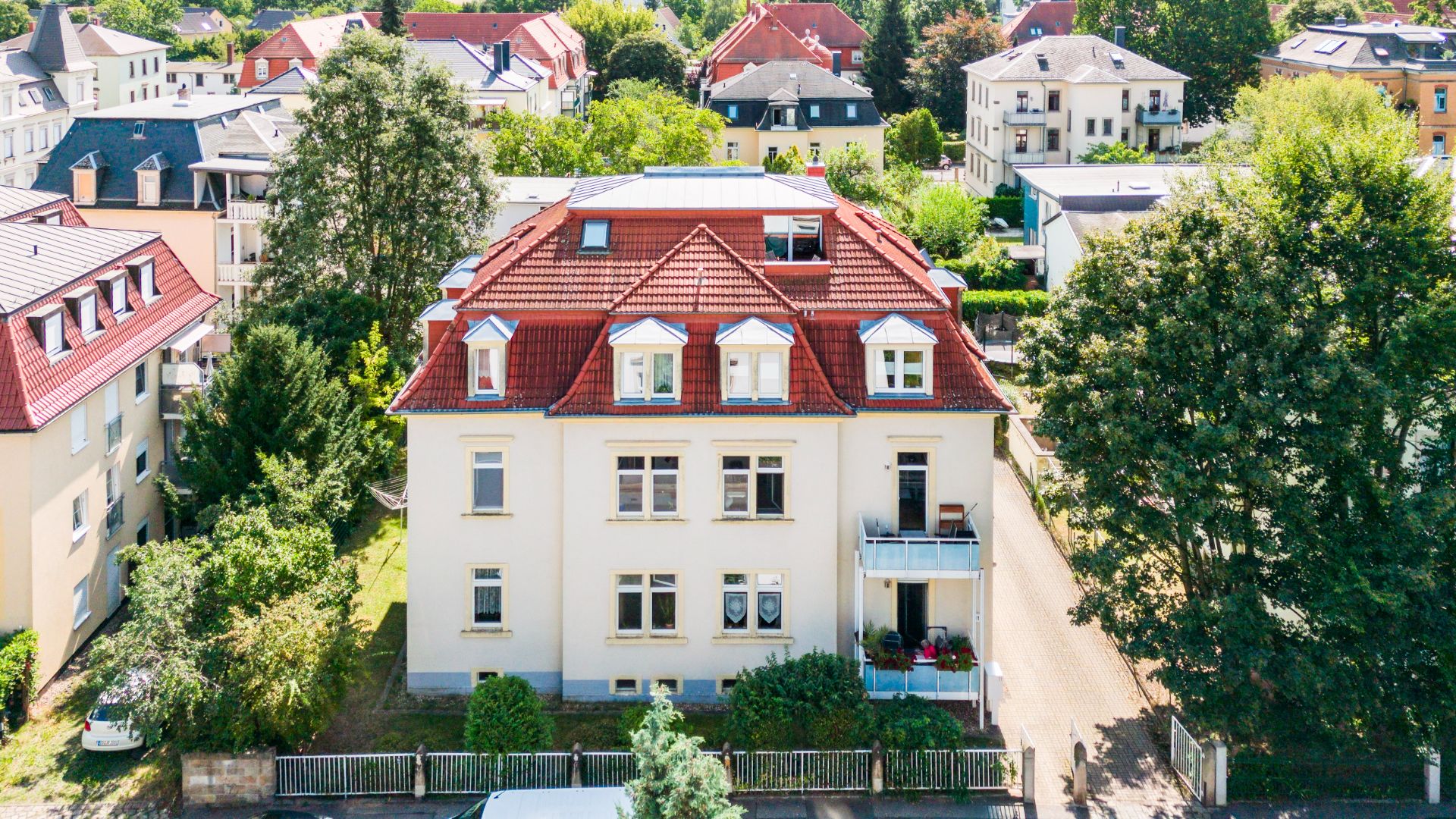 Dreistöckiges Wohnhaus mit rotem Dach, Balkon und Garten, umgeben von anderen Häusern und Bäumen.