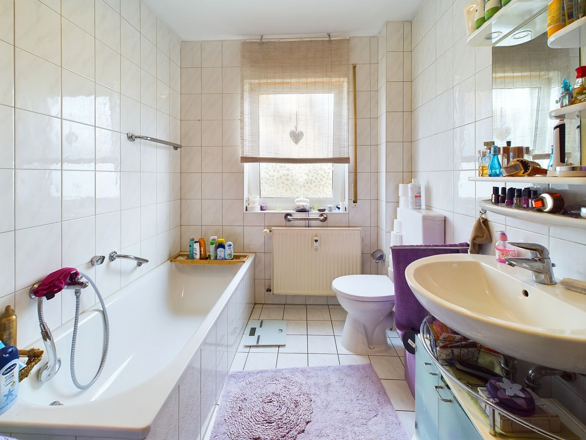 Helles Badezimmer mit Badewanne, Waschbecken, Toilette und Fenster, dazu Regale und lilafarbene Matte.