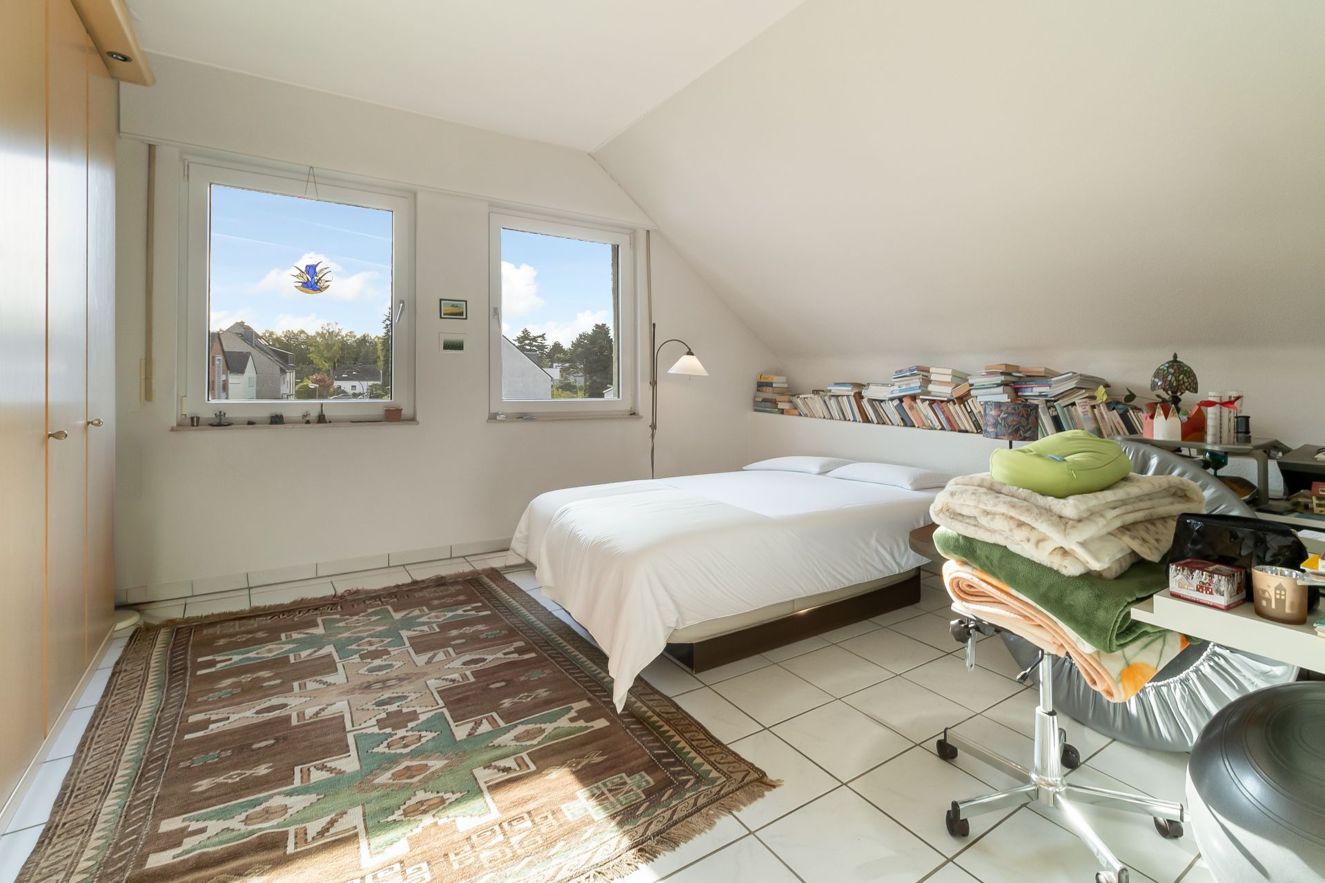 Helles Schlafzimmer mit Doppelbett, Teppich, Schreibtisch und Bücherregal unter schräger Decke.