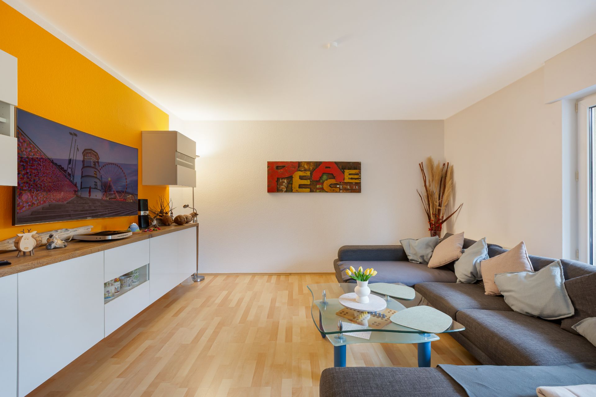 Helles Wohnzimmer mit grauem Ecksofa, Glastisch und gelber Wand.