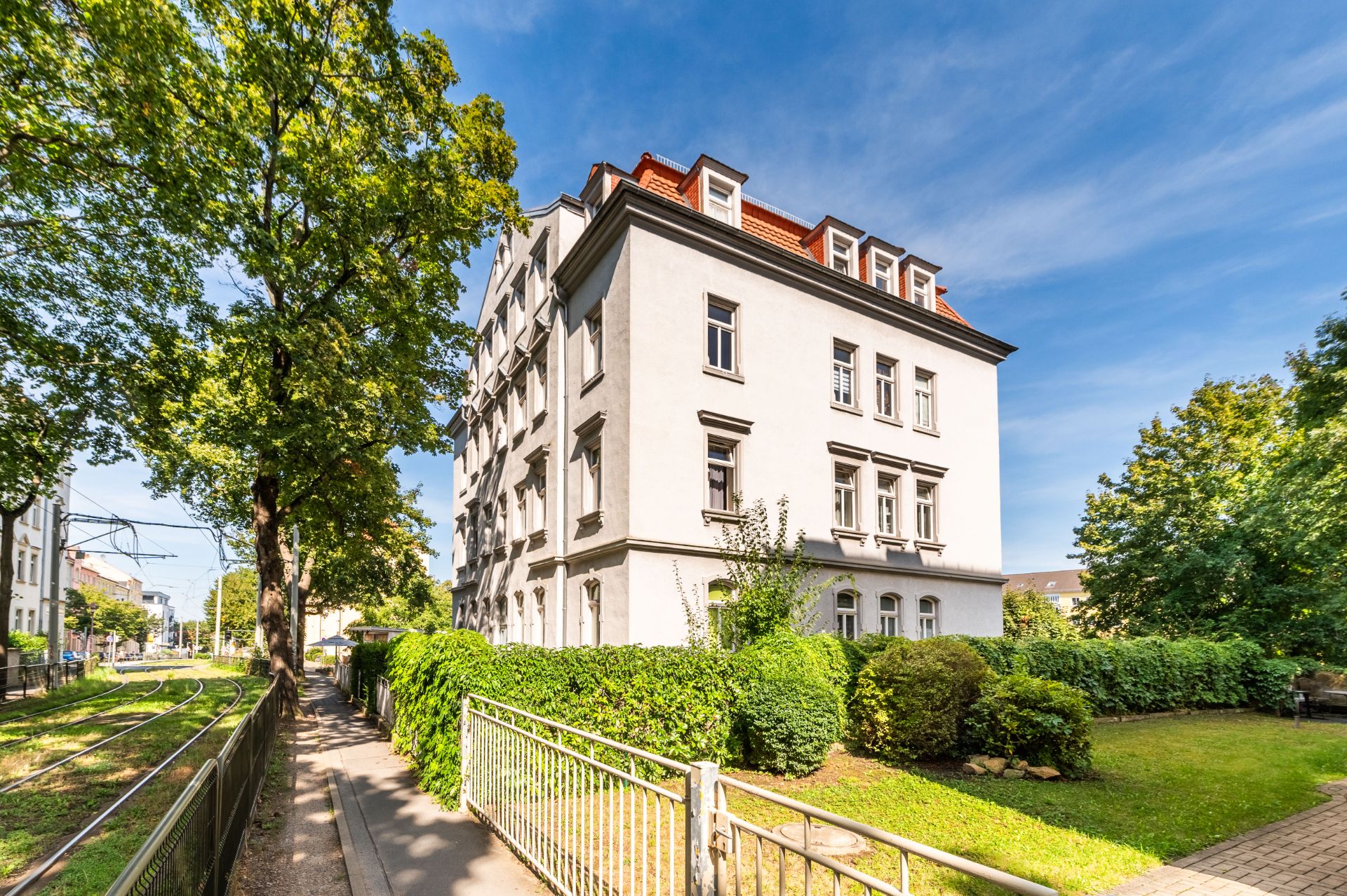 Mehrstöckiges Wohngebäude mit Garten und Gehweg neben einer Straßenbahnlinie.
