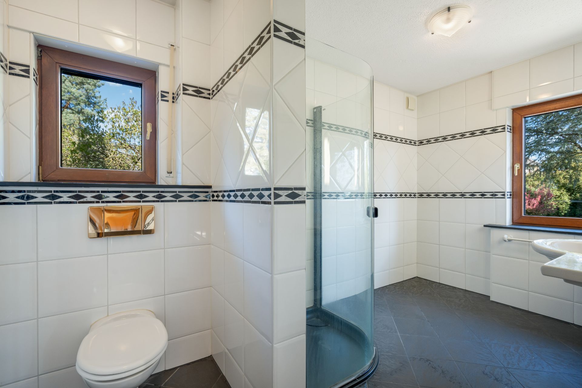 Modernes Badezimmer mit Toilette, Dusche, Waschbecken und zwei Fenstern, weiße Fliesen mit Dekorstreifen.