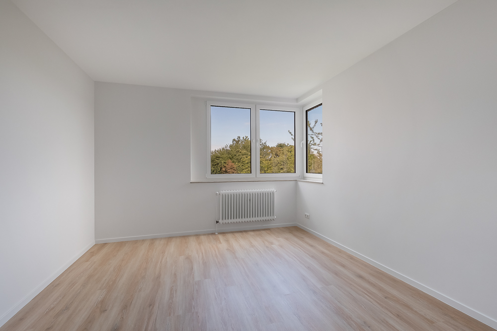 Leerer, heller Raum mit Holzfußboden und drei Fenstern mit Blick ins Grüne.