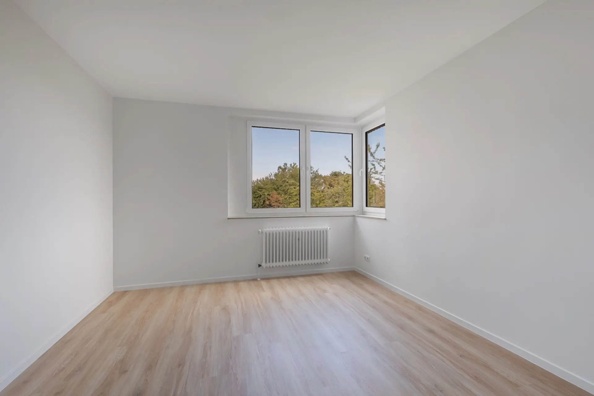 Leerer, heller Raum mit Holzfußboden und drei Fenstern mit Blick ins Grüne.