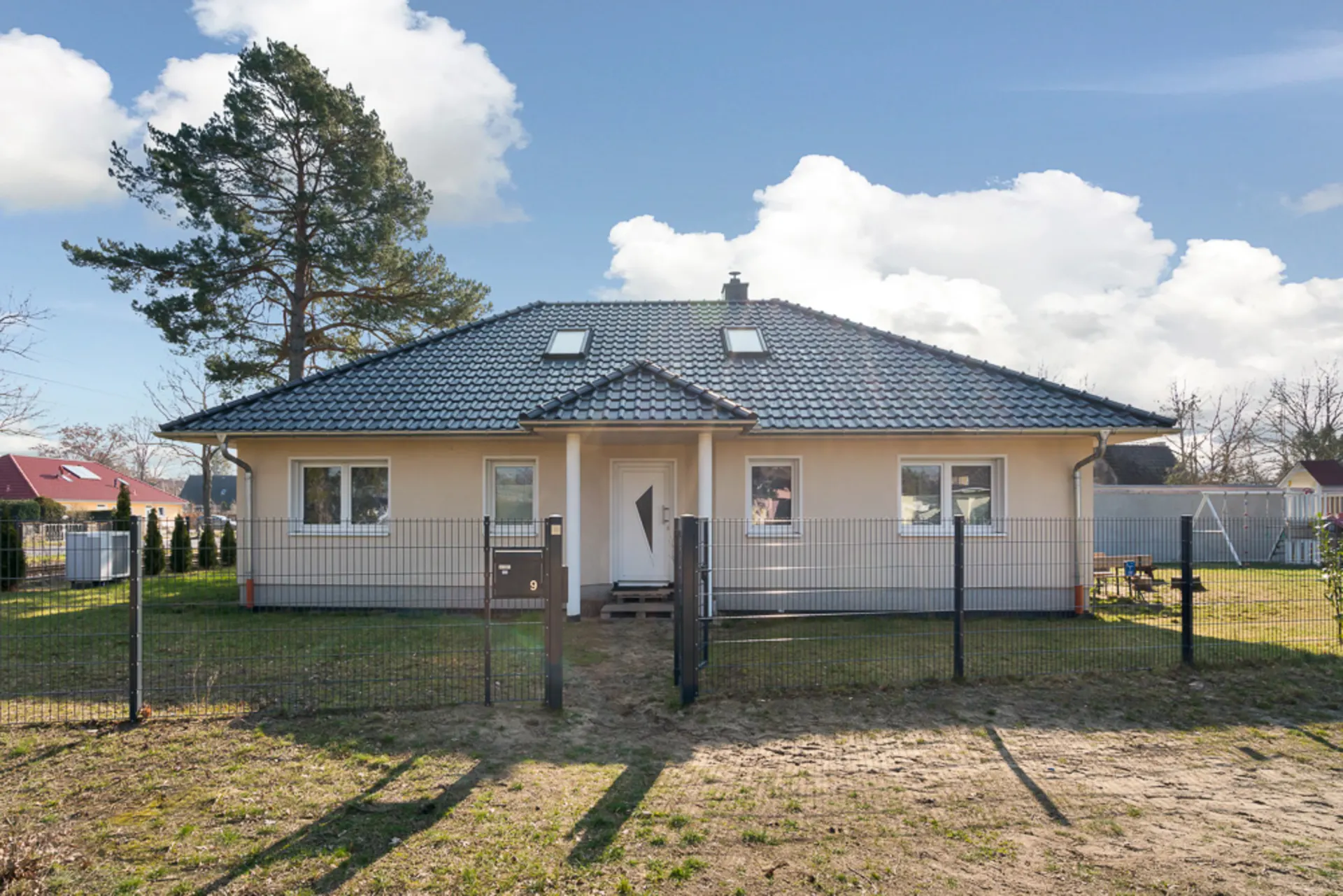 Einfamilienhaus mit dunklem Dach, hellem Putz und umzäuntem Vorgarten, frontal aufgenommen.
