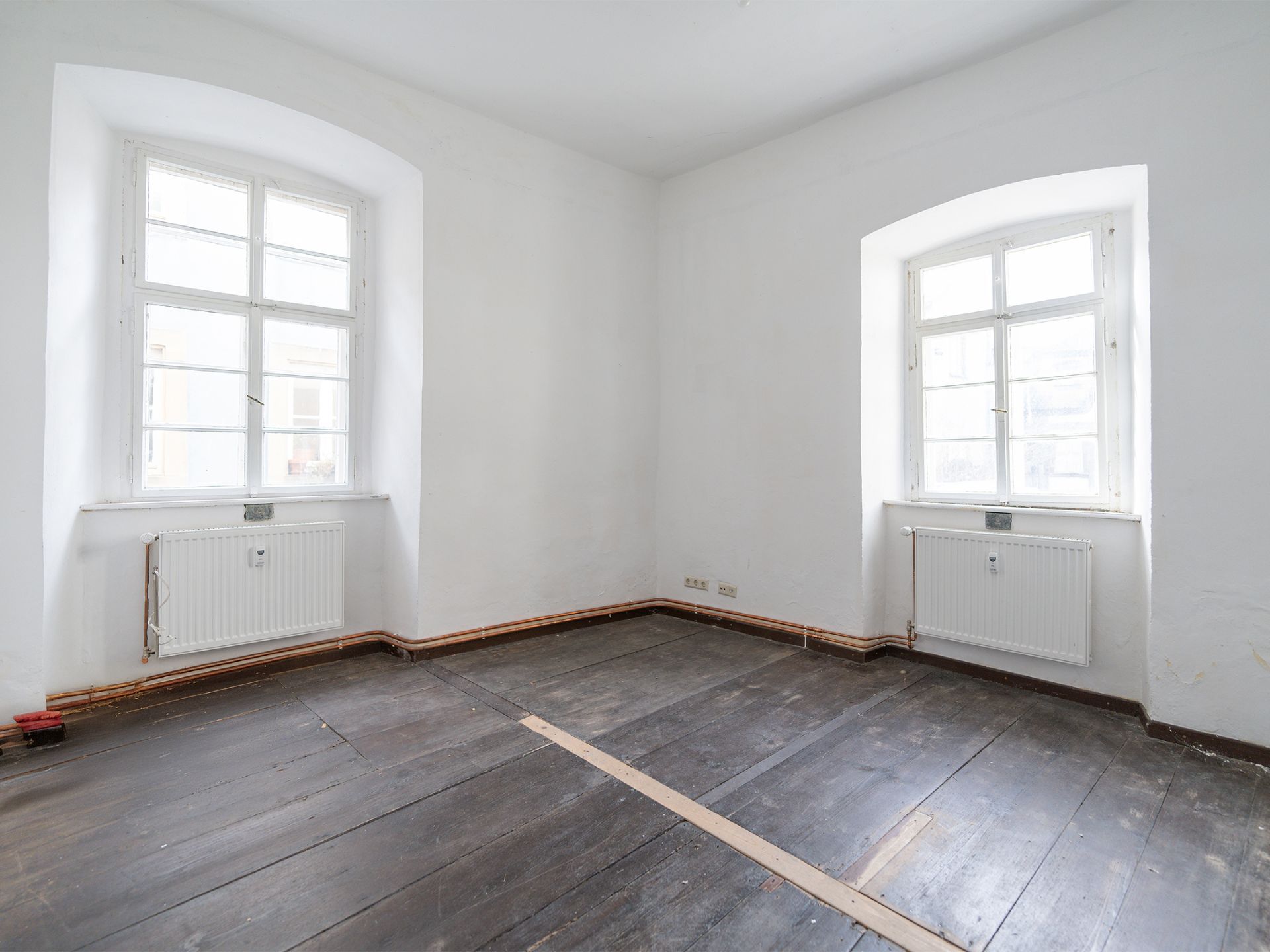 Leeres Zimmer mit zwei Fenstern, weißen Wänden und dunklem Holzboden.