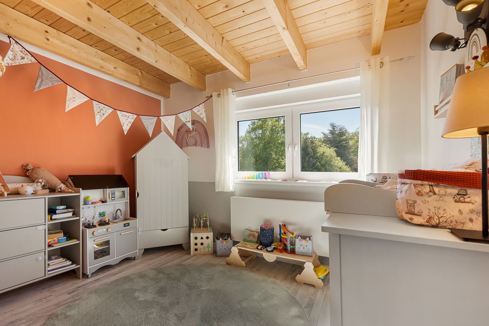 Helles Kinderzimmer mit Holzdecke, Spielküche, Teppich und Fenster mit Blick ins Grüne.
