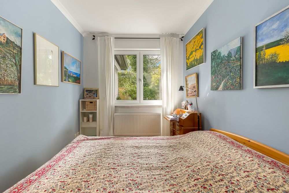 Schlafzimmer mit großem Bett, blauer Wandfarbe, Gemälden und Fenster mit weißen Vorhängen.