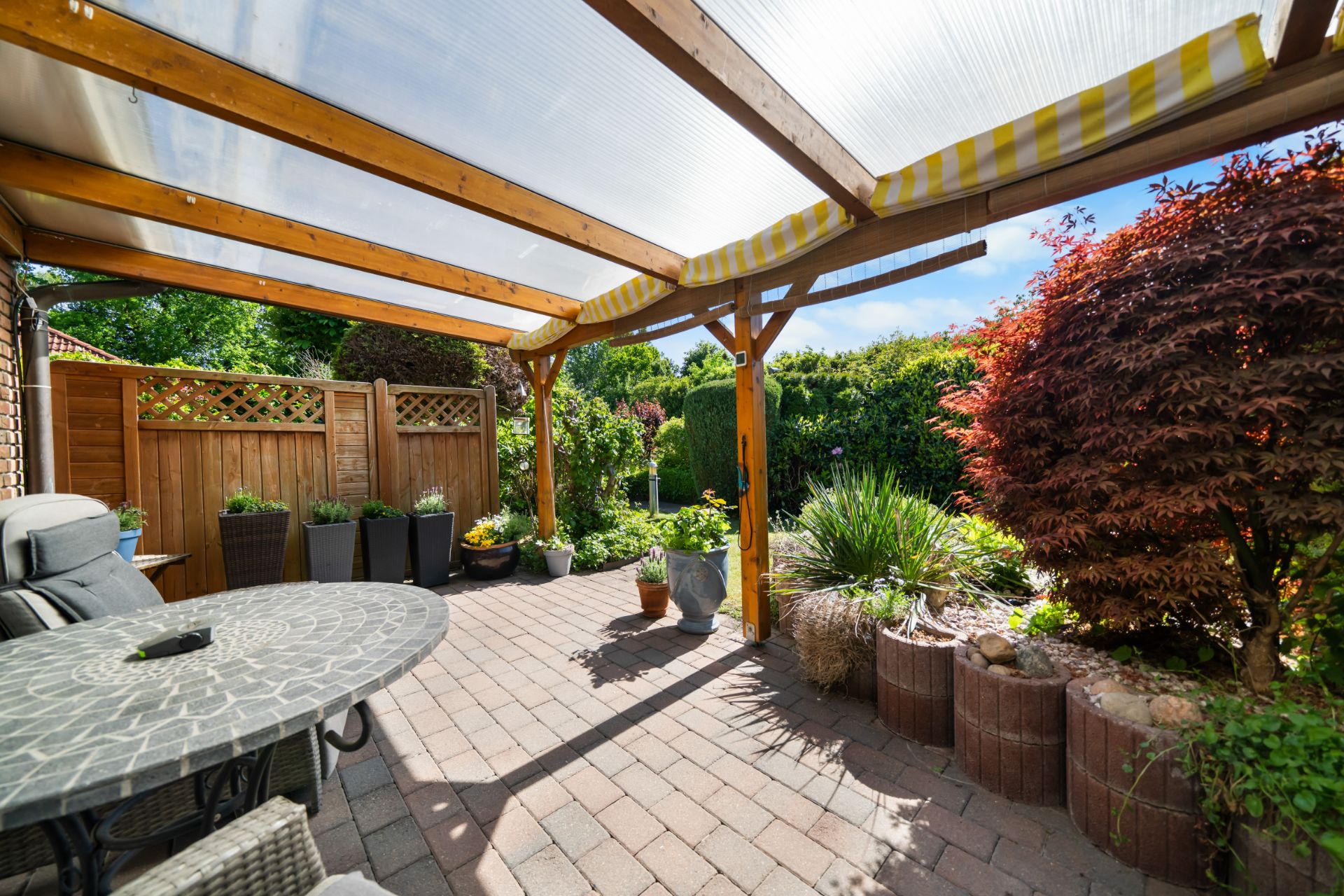 Überdachte Terrasse mit Sitzgruppe, Pflanzkübeln und Blick auf einen Garten.