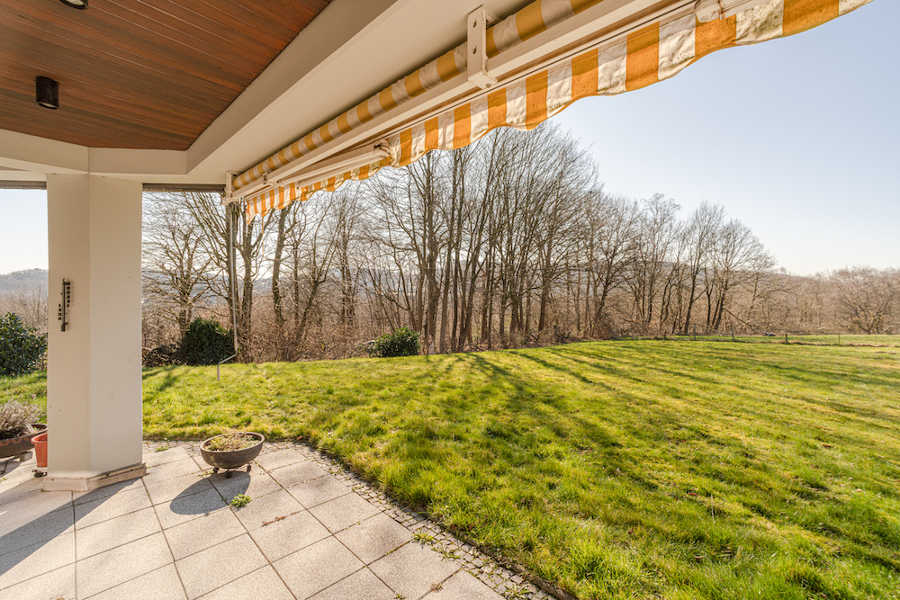 Überdachte Terrasse mit Markise und Blick auf einen großen Garten mit Rasen und Bäumen.