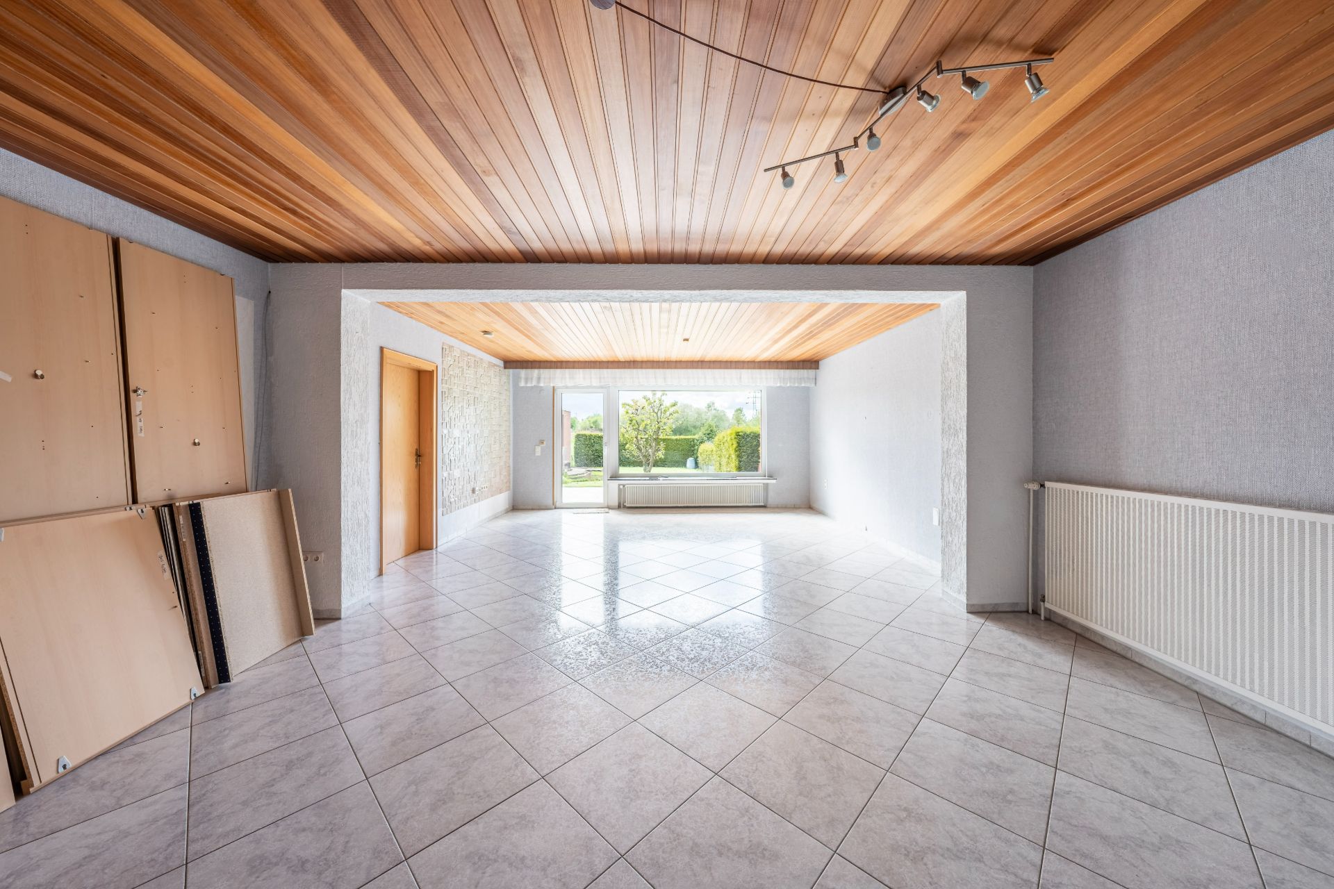 Heller leerer Raum mit Fliesenboden, Holzdecke, großem Fenster und ein paar an die Wand gelehnten Holzplatten.