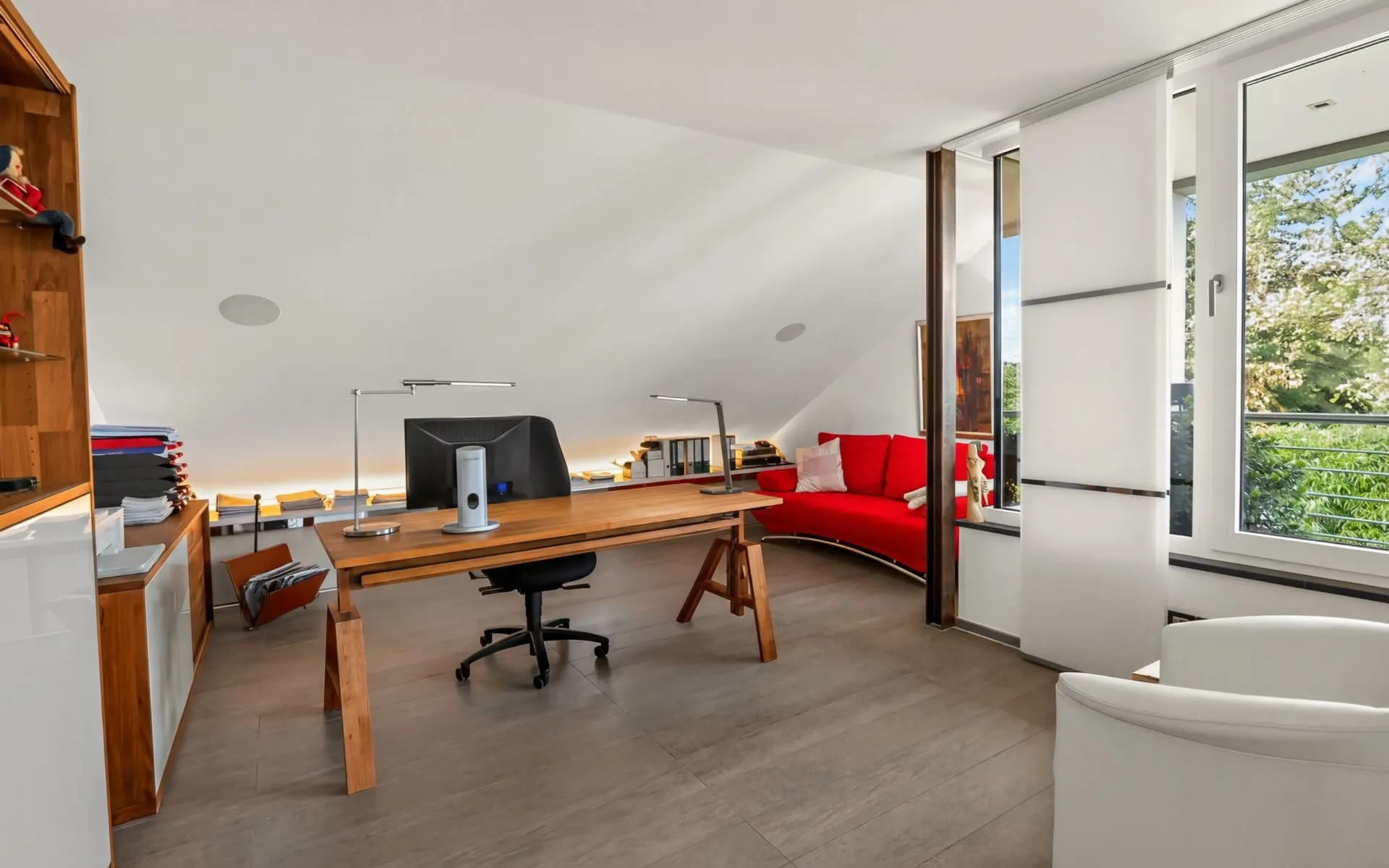 Helles Arbeitszimmer mit Holzschreibtisch, rotem Sofa, Regal und großem Fenster mit Blick ins Grüne.