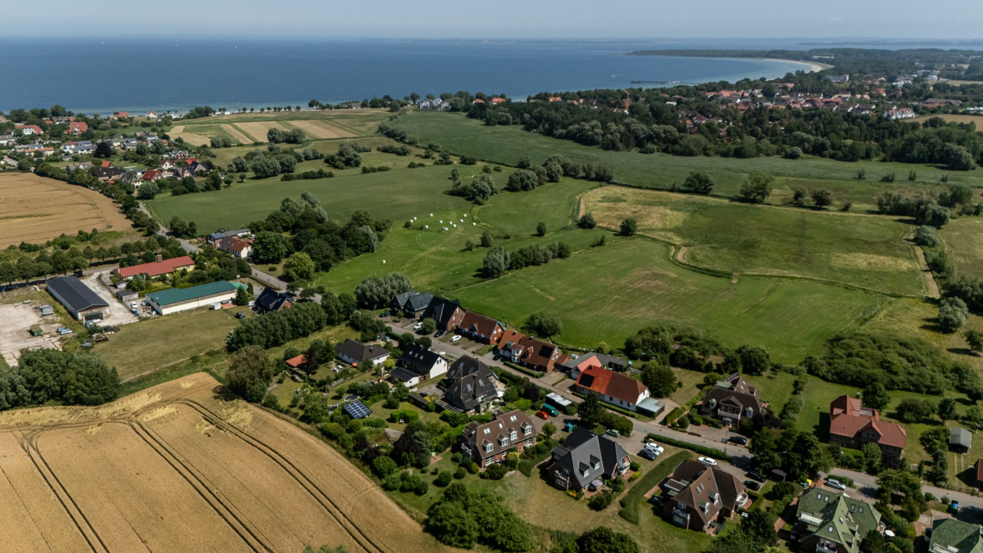 Luftaufnahme eines Wohngebiets mit Häusern, Feldern und Küste im Hintergrund.